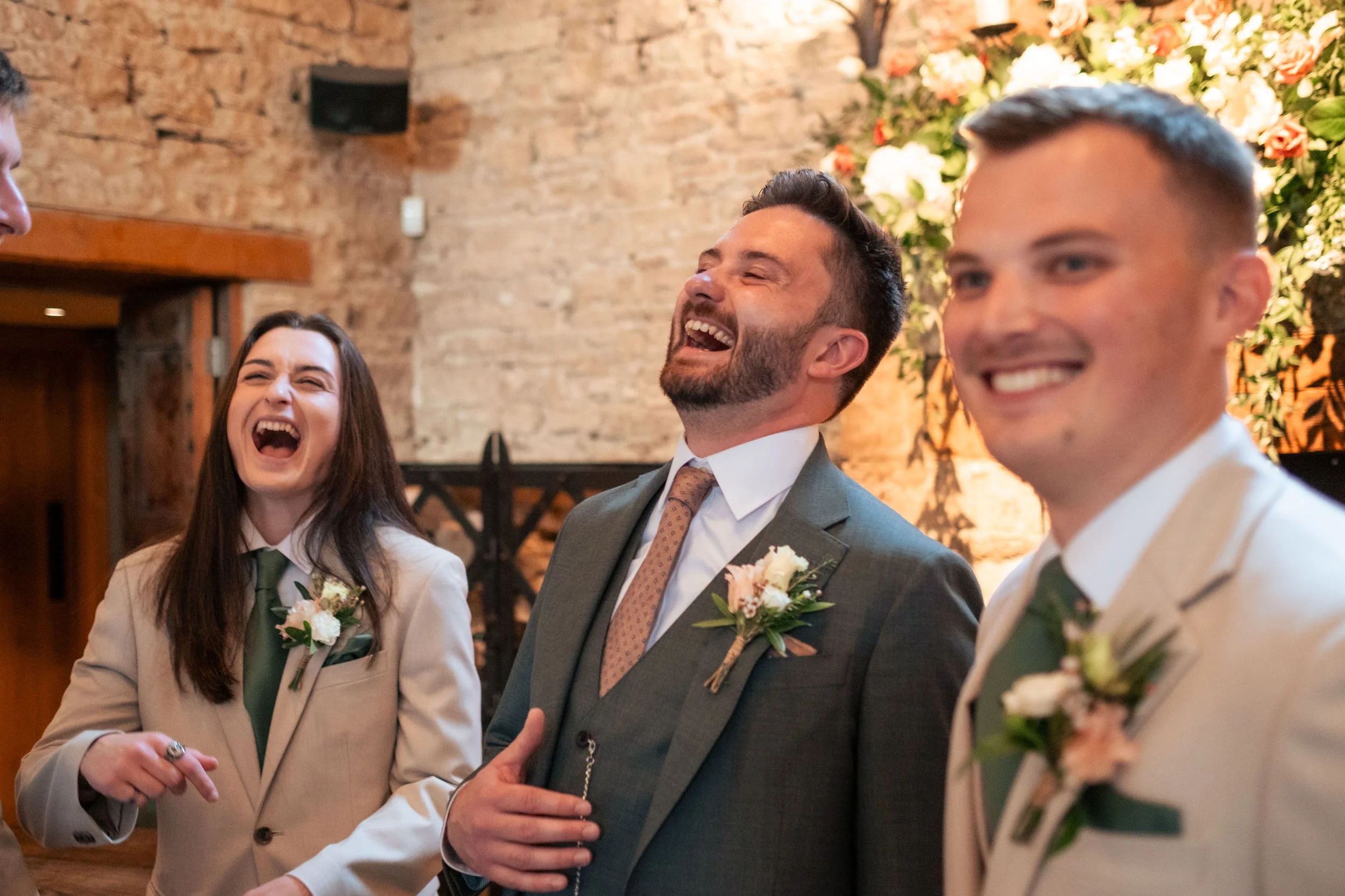 The groom and his grooms-people laughing and enjoying themselves before the wedding inside a rustic venue with brick walls and floral decorations.