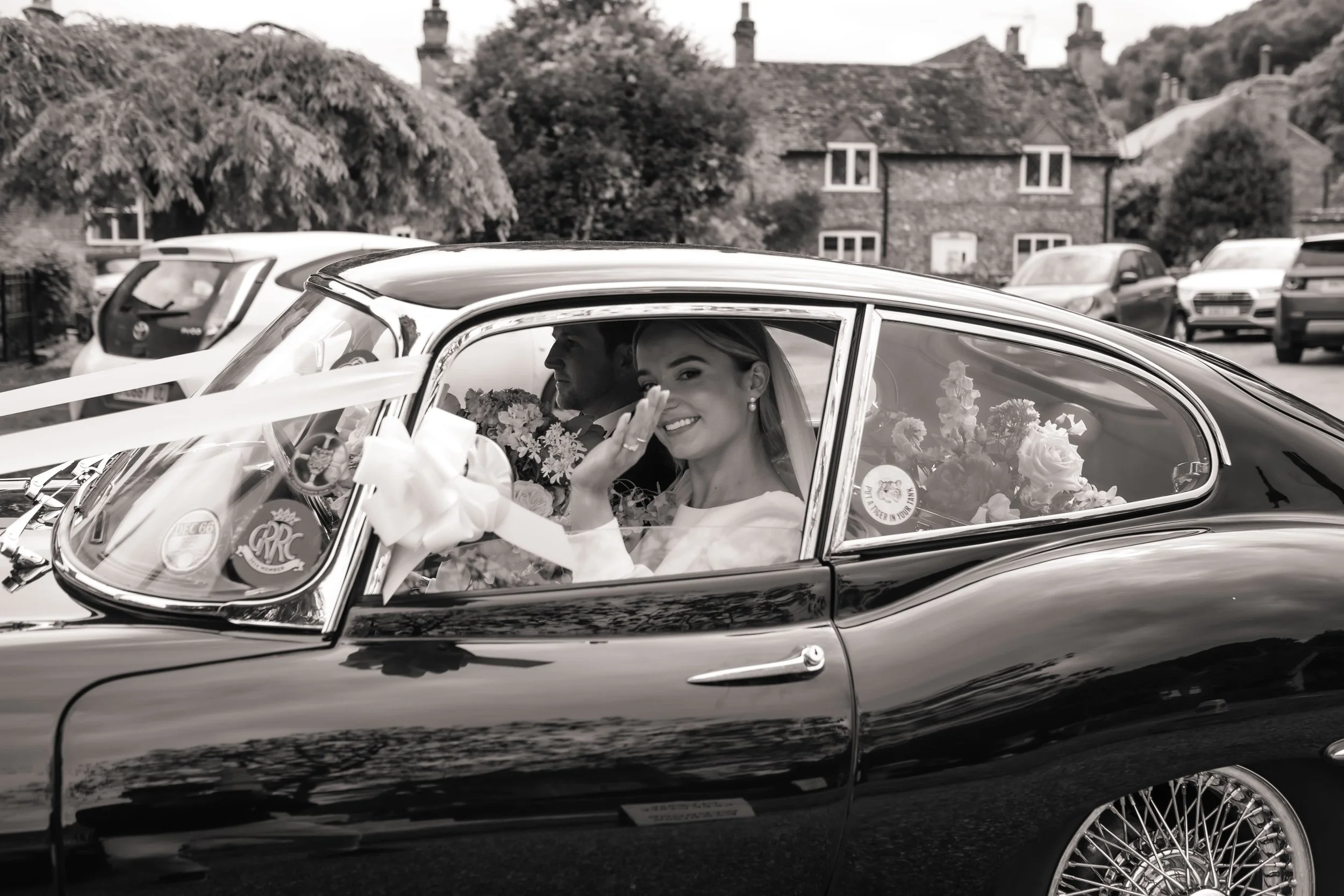 A bride smiling and waving from inside a vintage black car decorated with flowers, with a wedding dress and veil visible. The groom is sitting next to her, holding a bouquet, in Hambleden, Berkshire.