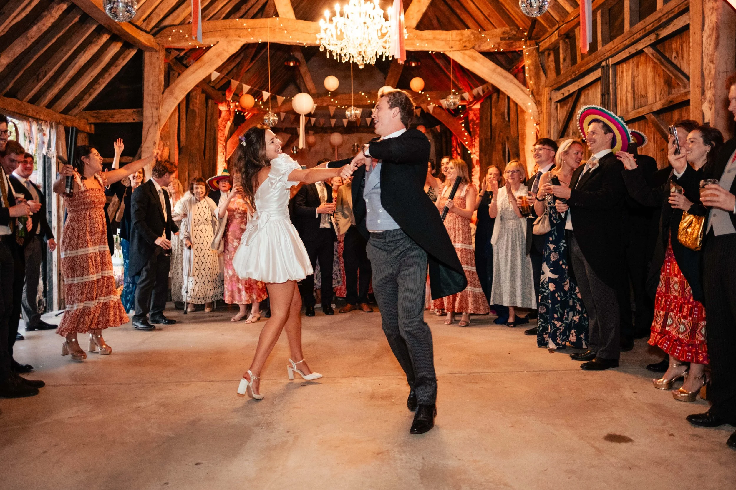 A couple dancing at a wedding reception with guests surrounding them in a rustic barn decorated with string lights and paper lanterns.