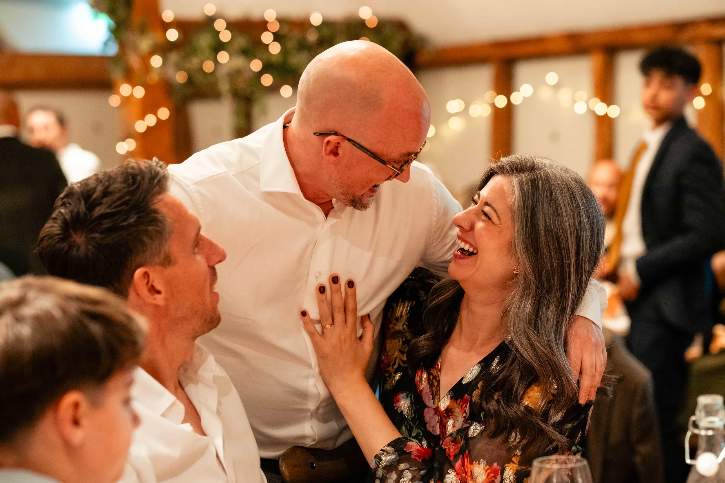 Happy woman and middle-aged man embracing at a celebration event, surrounded by friends, with warm lighting and festive decor in the background.