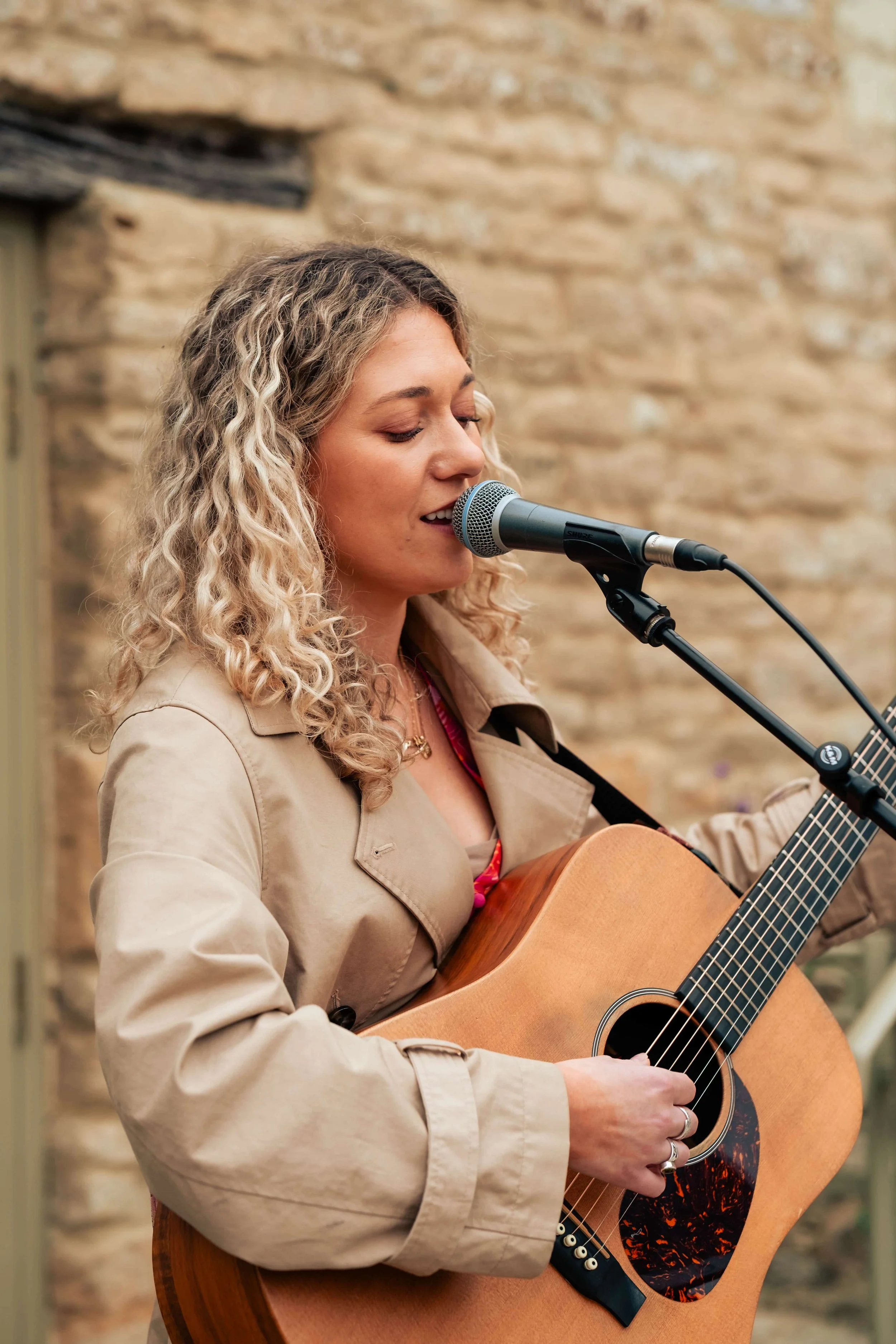 Woman singing into a microphone while playing an acoustic guitar outdoors against a stone wall.