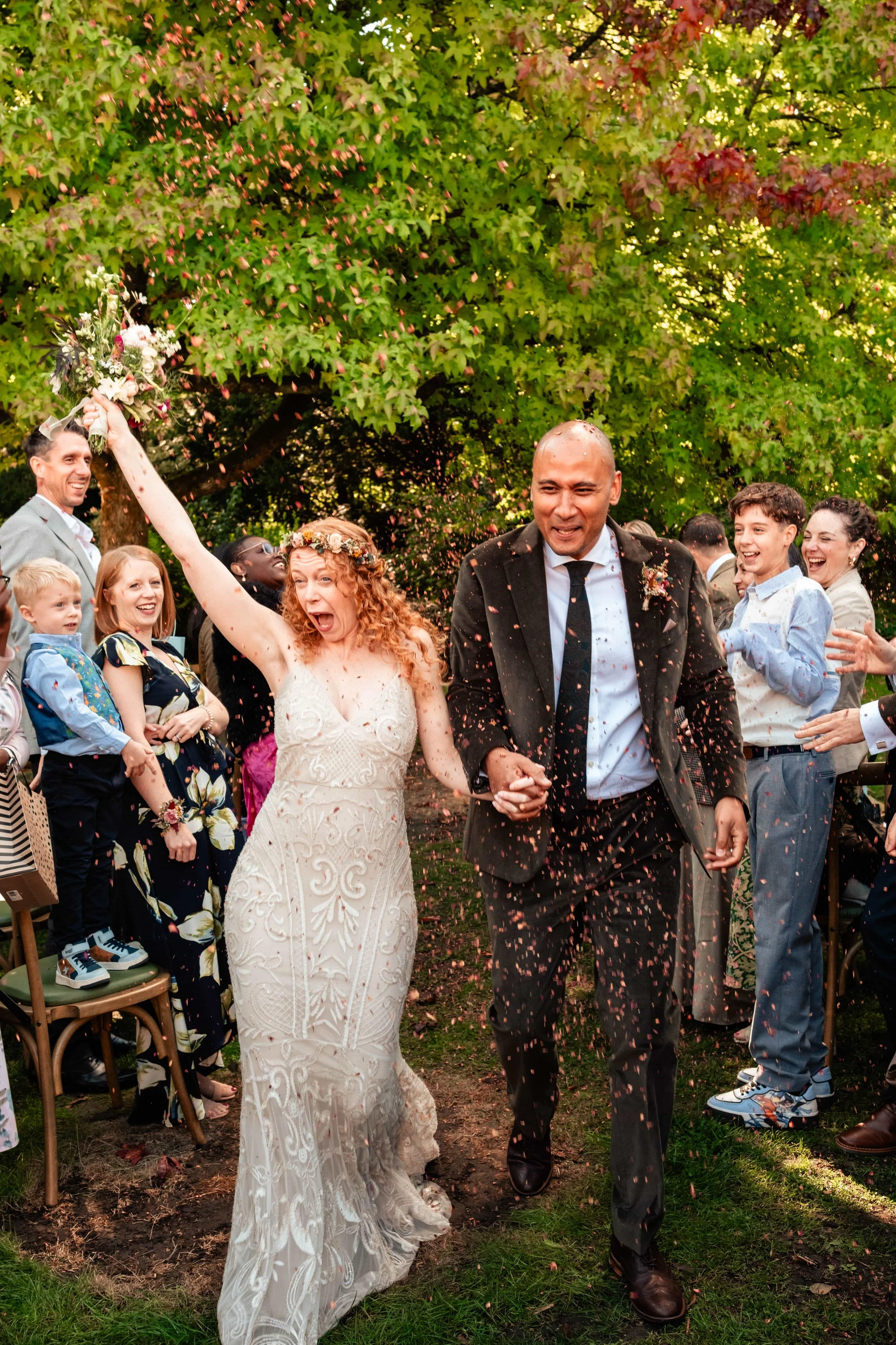 Bride and groom joyful walking through a celebration, surrounded by cheering guests outdoors near green trees.