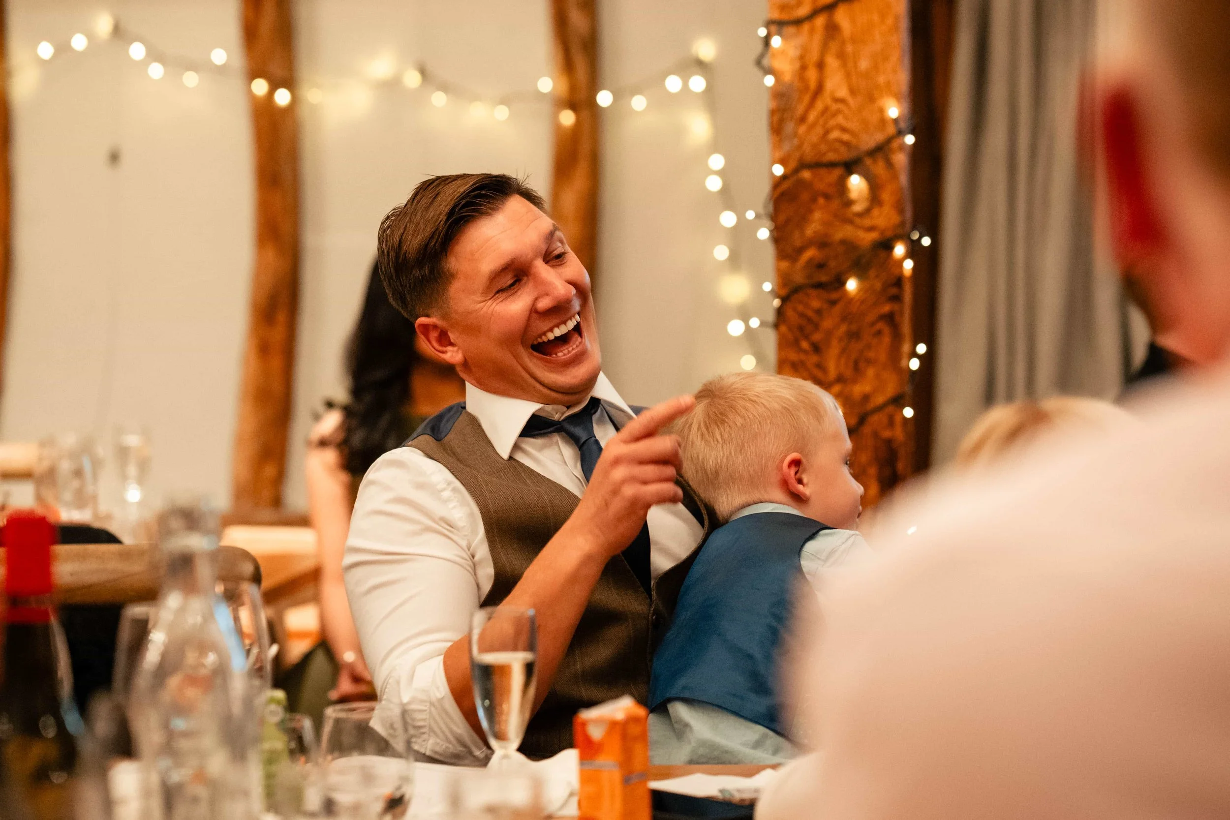 A man with a wide smile, wearing a vest and tie, sitting at a table with a young boy leaning on him. The scene takes place in a warmly lit room decorated with string lights and wooden beams, likely at a celebration or party.