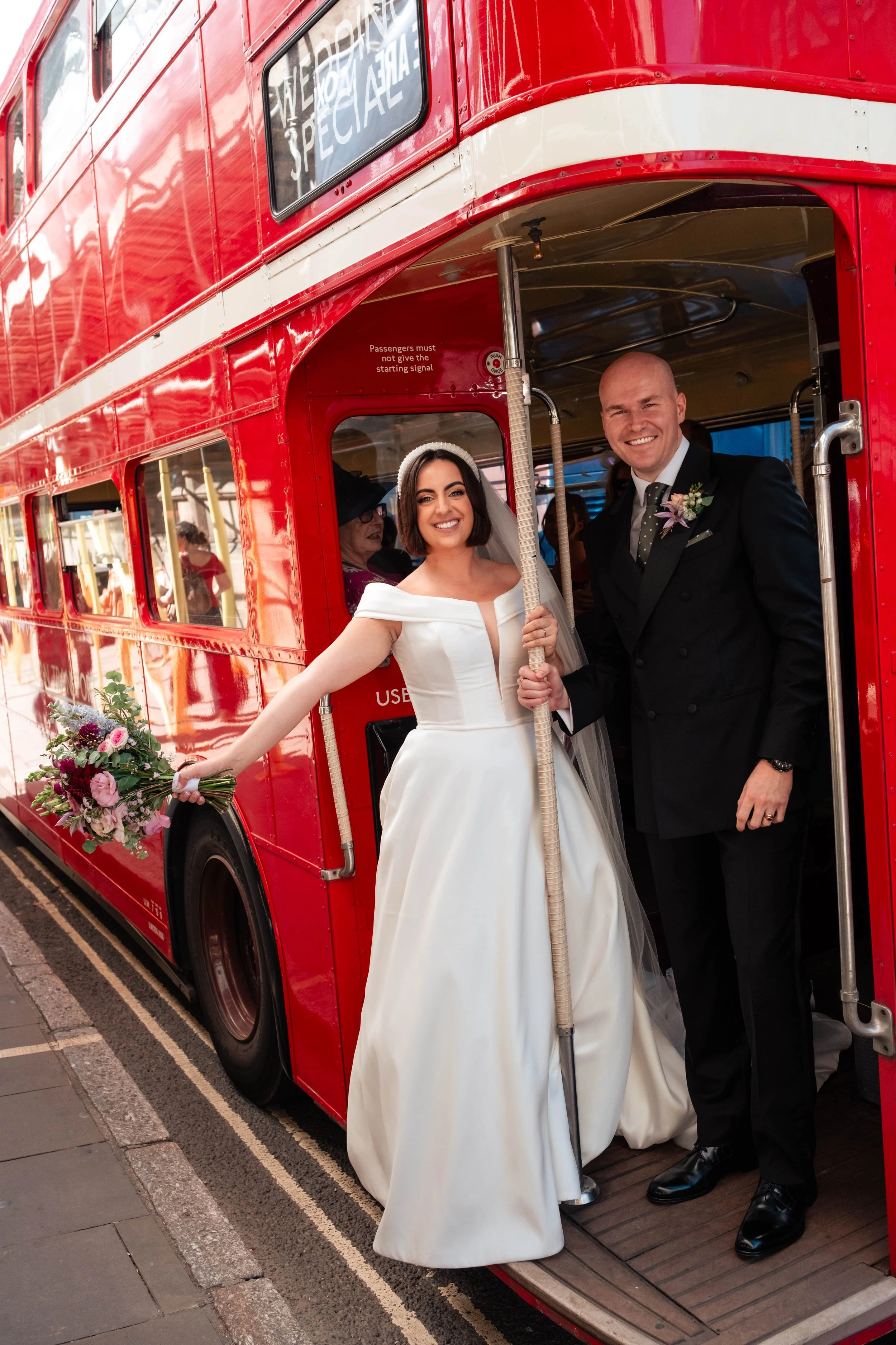 Bride and groom standing on the lower deck of a red double-decker bus, smiling for a photo, with the bride holding a bouquet of flowers and the groom dressed in a black suit with a boutonnière.
