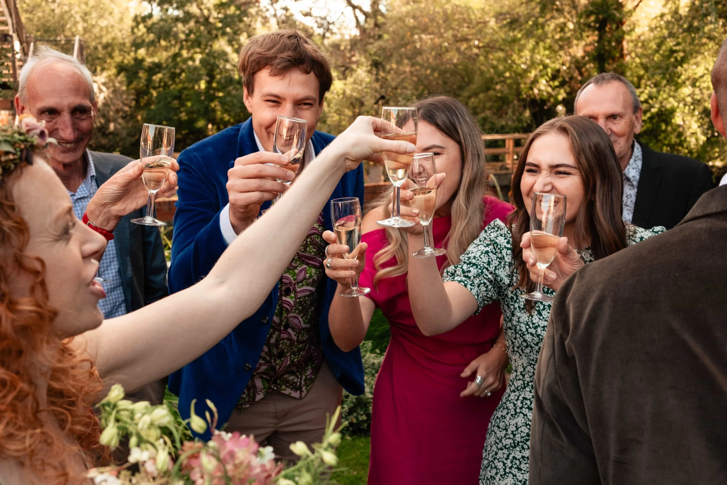 Group of people raising glasses for a toast at an outdoor celebration, smiling and enjoying each other's company in a garden setting.