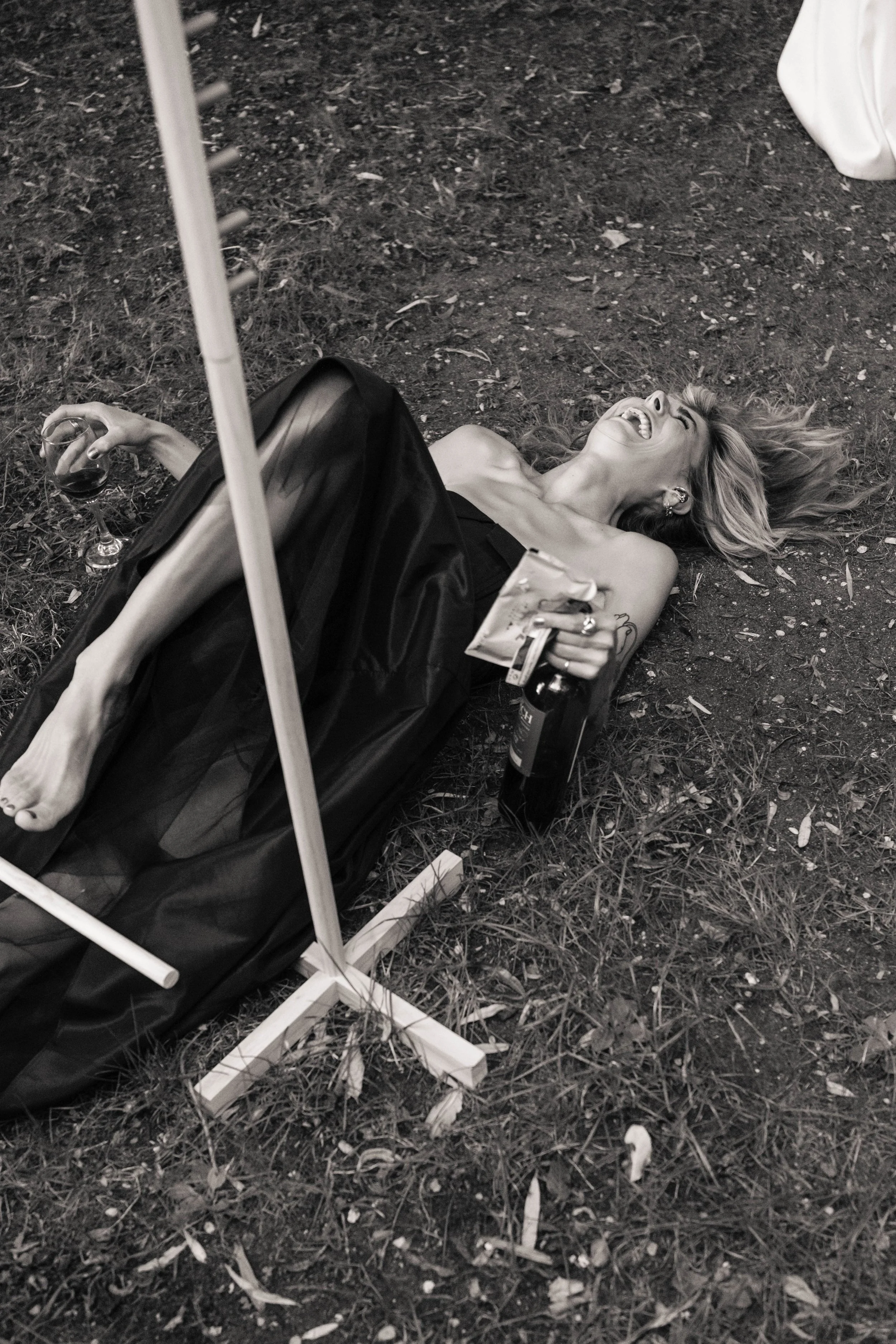 A woman lying on the ground outdoors, laughing with her head tilted back, holding a glass of wine and a book or menu, with a bottle of wine nearby, under a stand or umbrella.