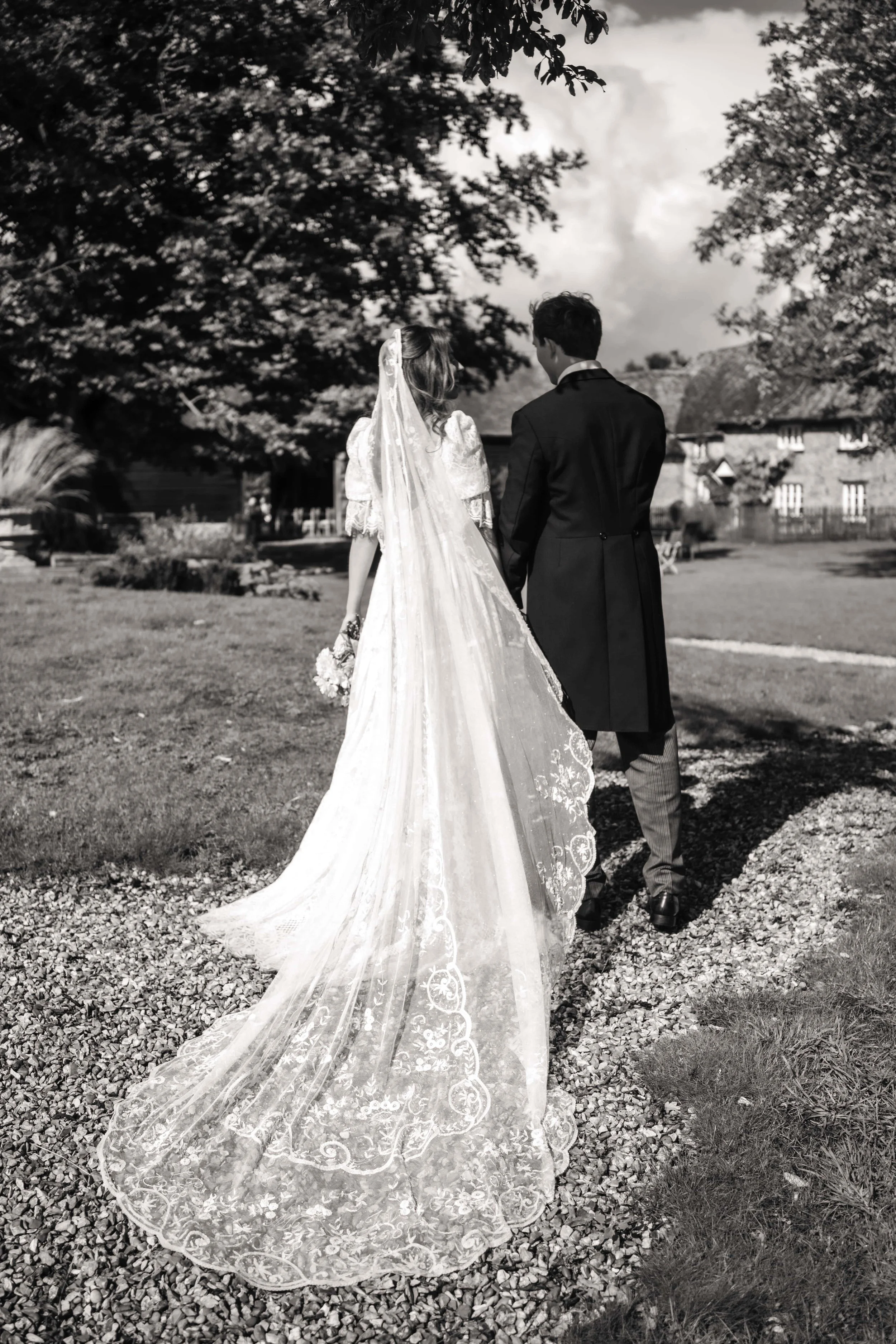 A black and white photograph of a bride and groom walking away on a gravel path in a garden, with the bride wearing a long, lace wedding dress and the groom dressed in a formal coat, holding hands.