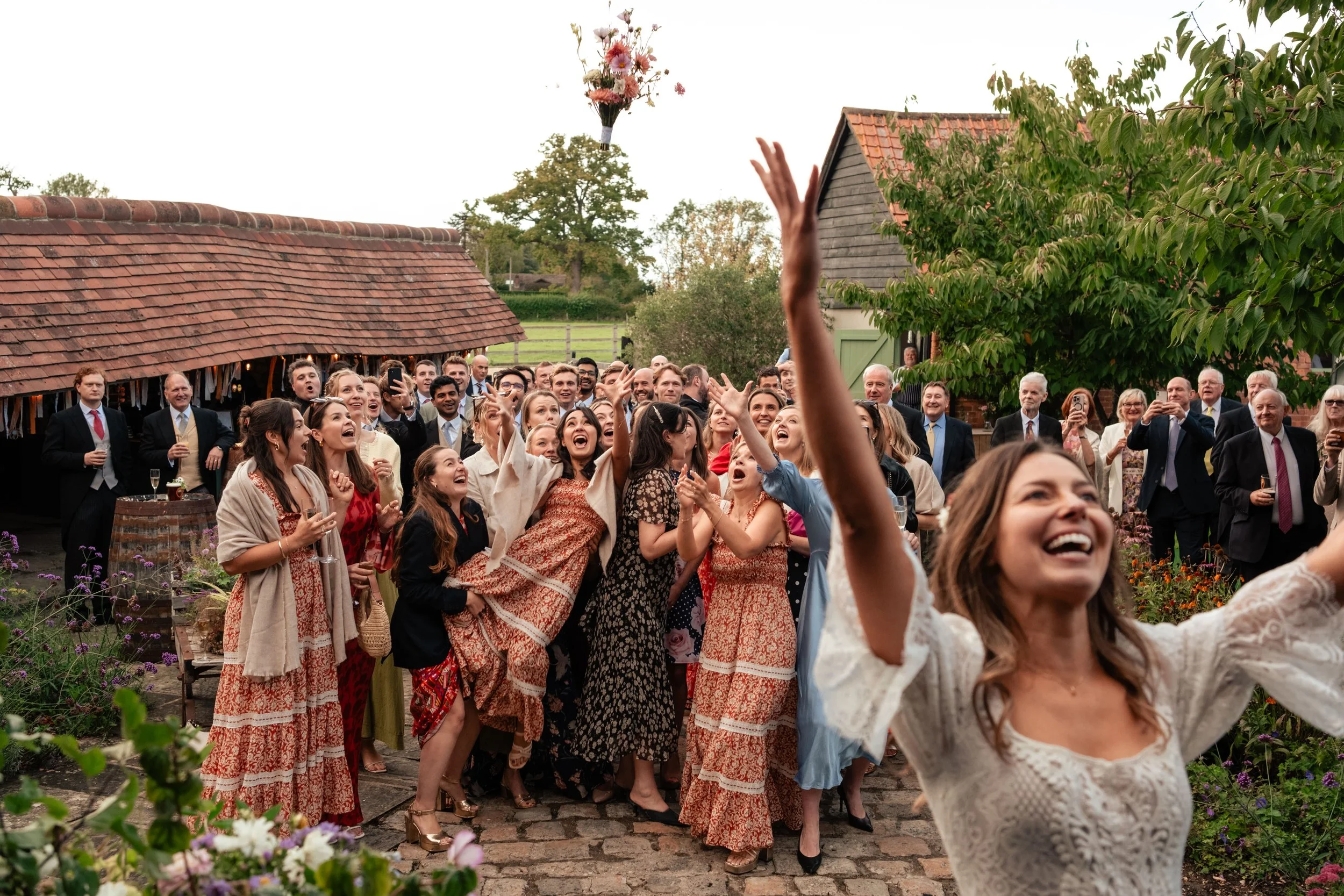Guests celebrating at an outdoor wedding reception as the bride smiles and throws her bouquet for the crowd to catch.