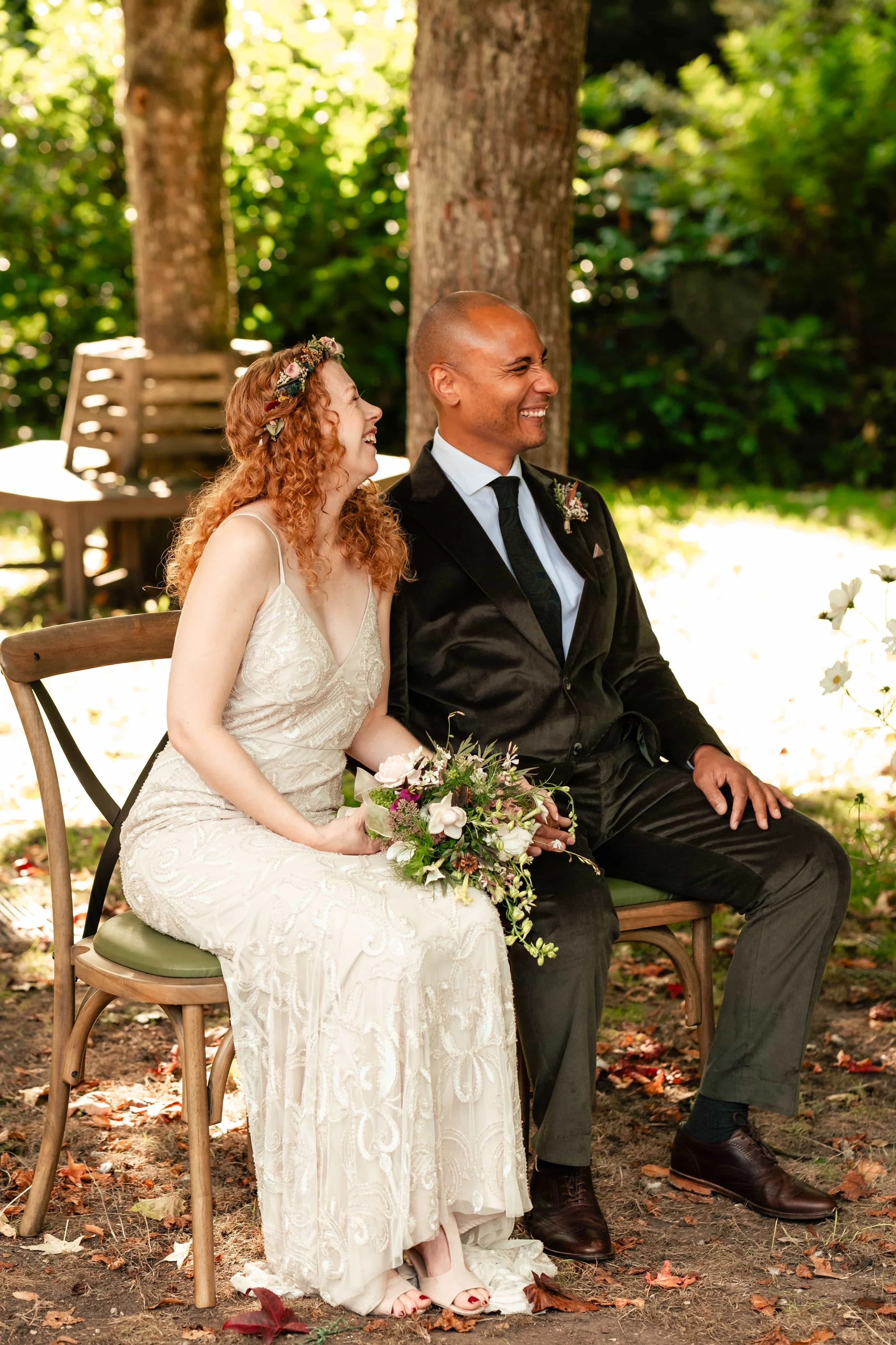 A couple sitting outdoors on a bench, dressed in wedding attire, smiling and sharing a joyful moment during their wedding ceremony surrounded by trees and greenery.