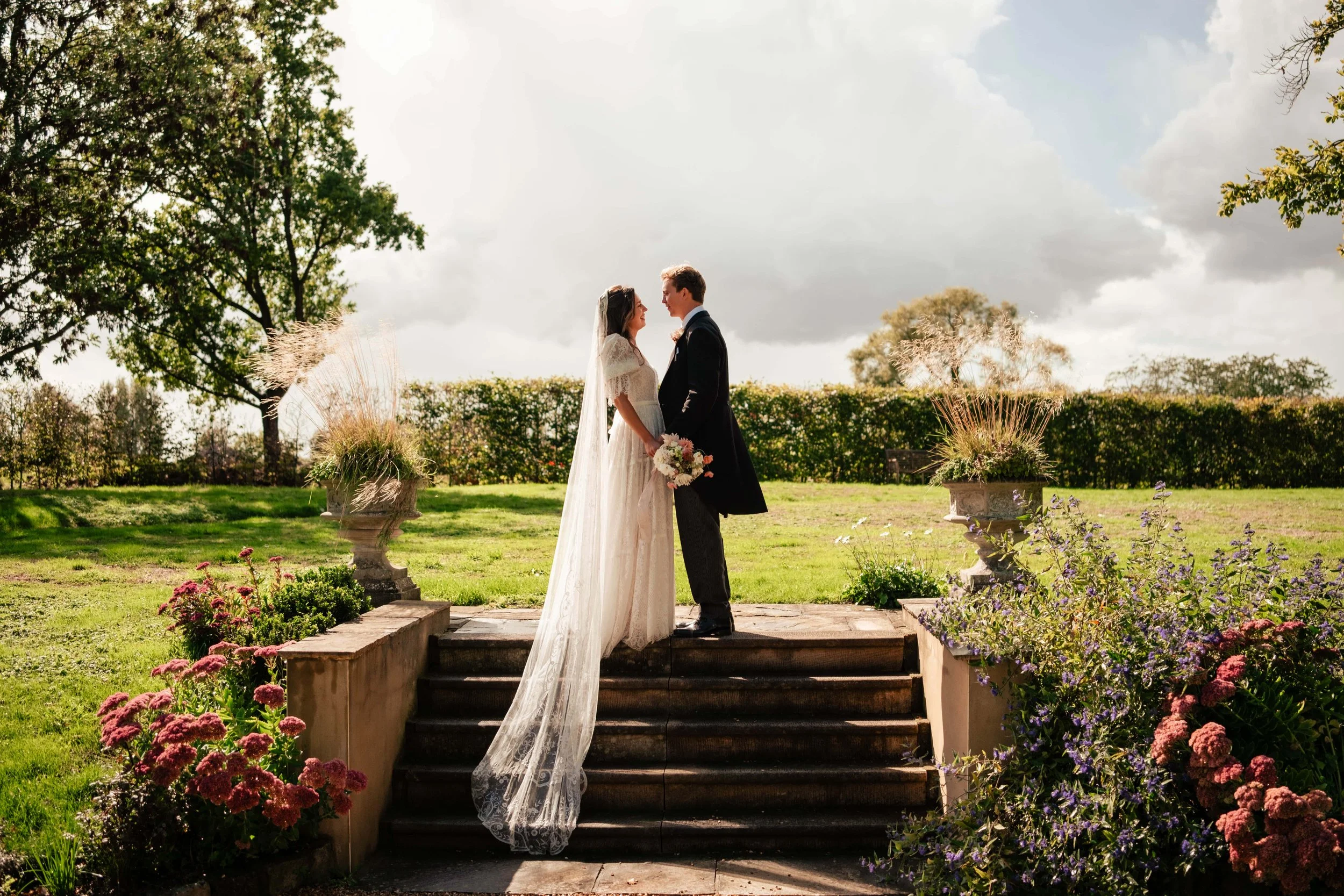 A bride and groom standing on the steps of a garden altar, holding hands and facing each other, with greenery and flowers around them and a cloudy sky in the background.