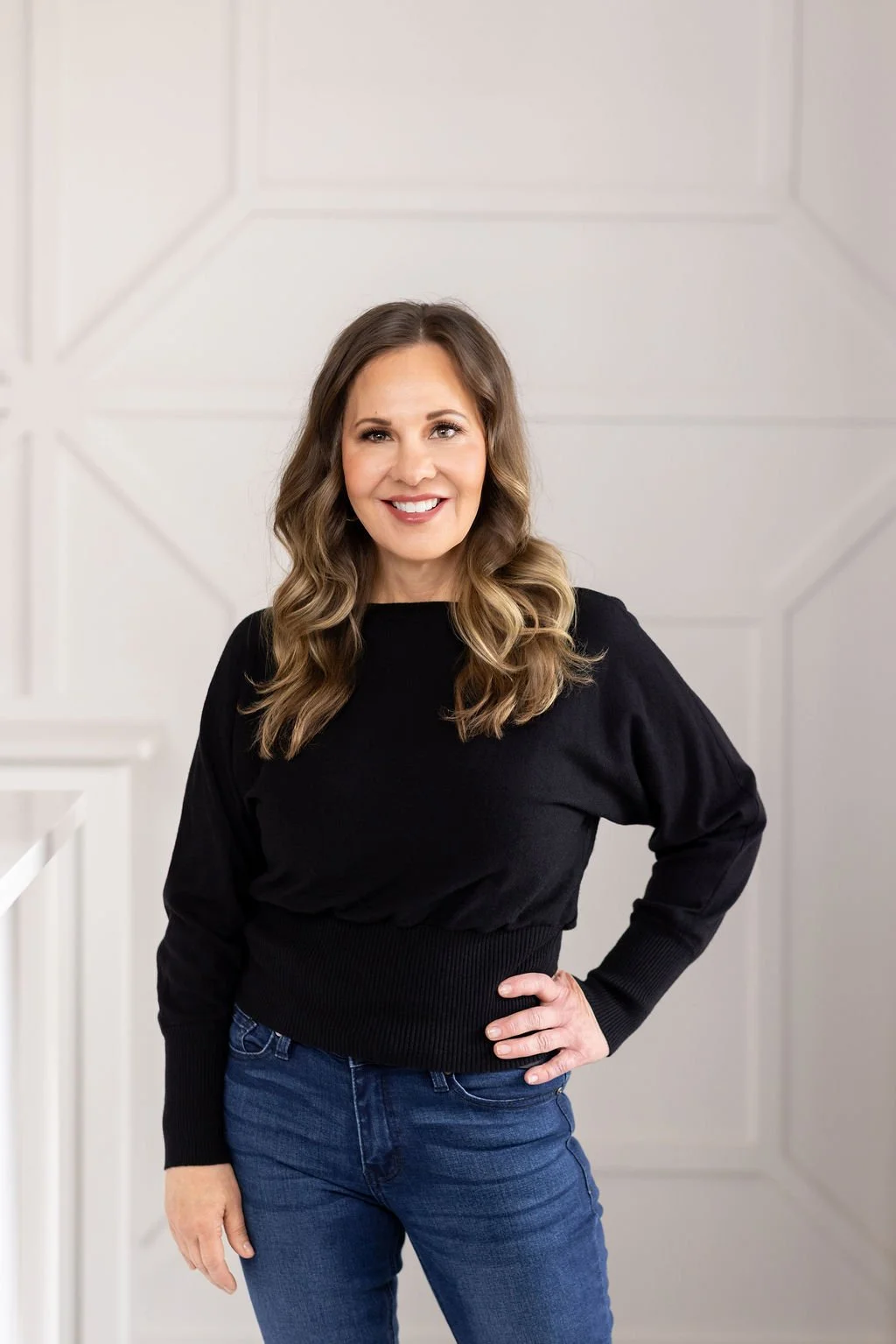 A woman with wavy, shoulder-length hair smiling in front of a beige tufted headboard.