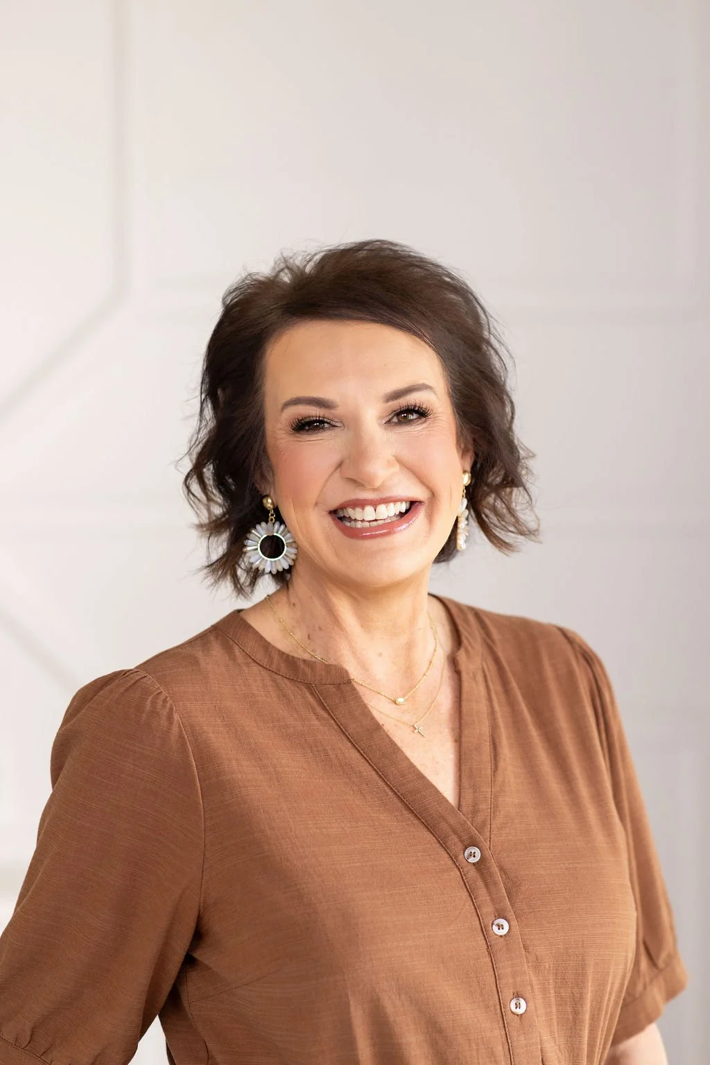 A middle-aged woman with shoulder-length brown curly hair, wearing gold hoop earrings, a delicate necklace with a small pendant, and a black top, smiling while sitting on a beige tufted sofa in front of a plain wall.