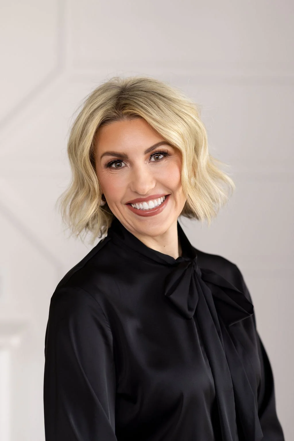 A woman with blonde, wavy hair smiles at the camera, wearing makeup and a white jacket, in front of a beige tufted background.