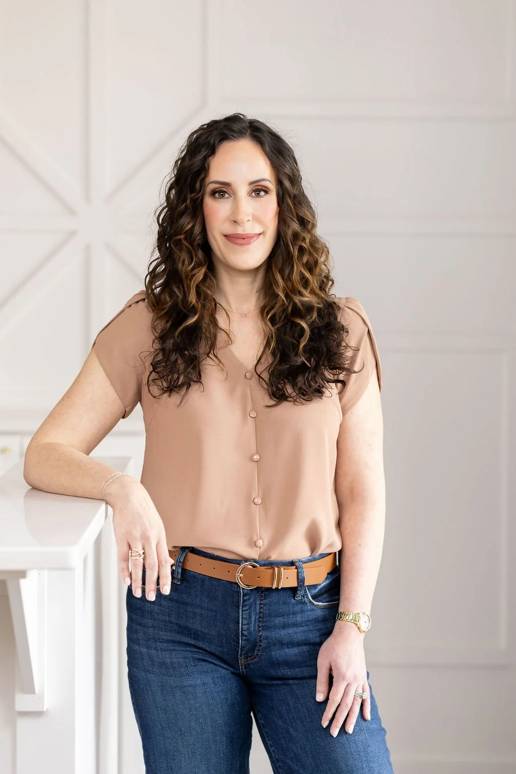 A woman with long curly brown hair smiling, wearing a maroon top and a delicate necklace, standing in front of a textured light-colored background.