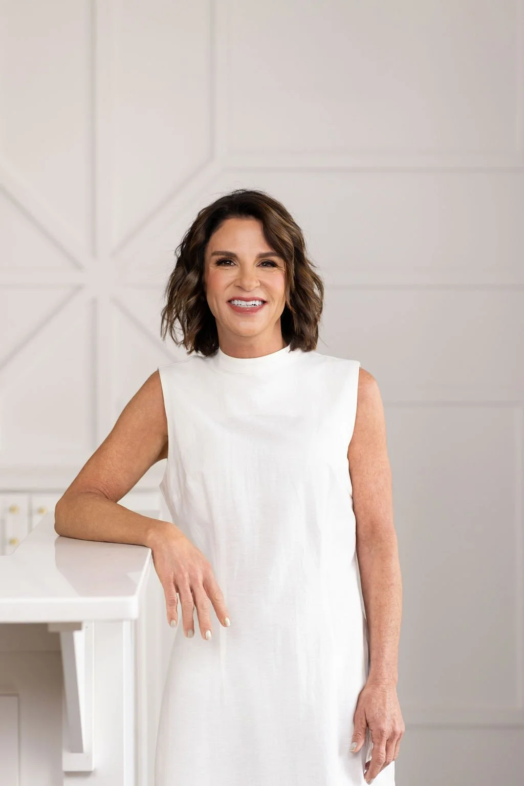 A woman with short, dark, wavy hair smiling at the camera. She is wearing statement earrings and a black top, sitting on a beige tufted chair in an indoor setting.