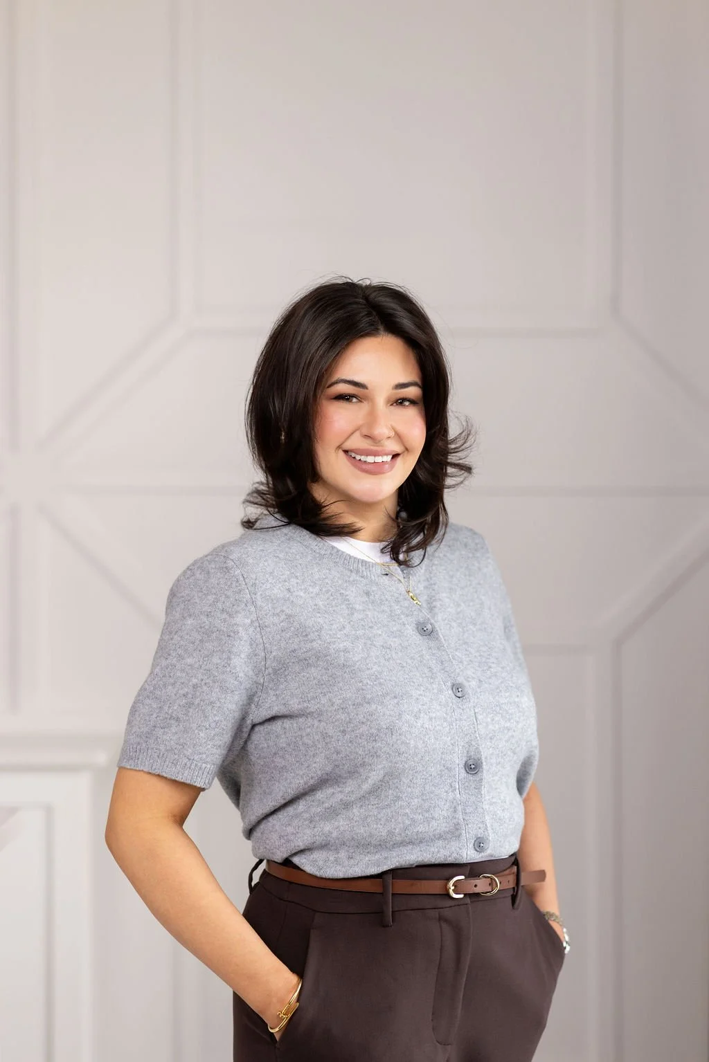 Portrait of a smiling woman with dark, wavy hair, wearing a light-colored blazer, jewelry, and makeup, sitting on a beige tufted headboard, in an indoor setting.