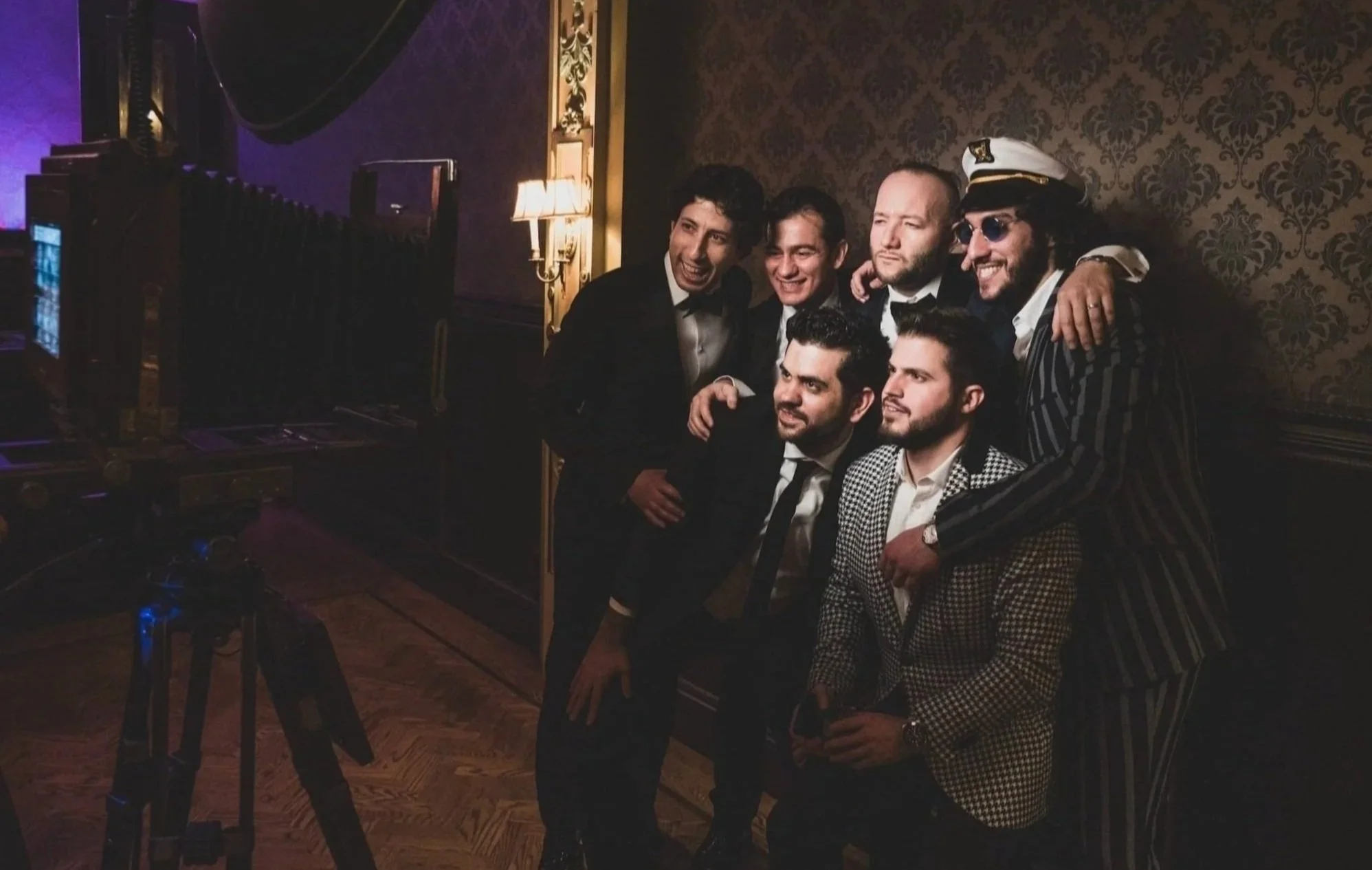 Group of six men dressed in formal and stylish outfits, posing for with a photo booth at a party or event in a dimly lit room with ornate wallpaper and wood flooring.
