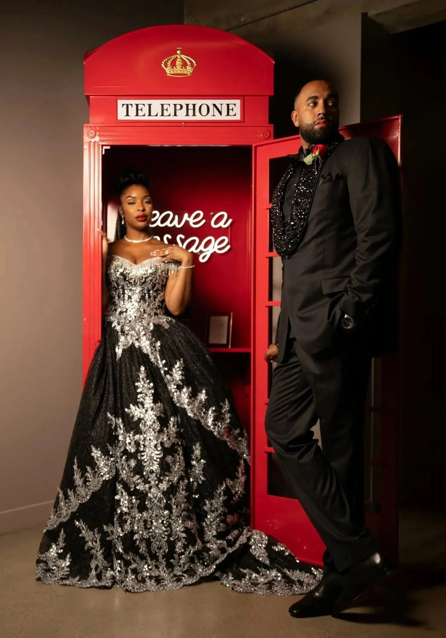 A woman in a black and silver glittery gown standing next to a man in a black tuxedo, both posing near a red telephone booth with a neon sign reading 'leave a message' inside.