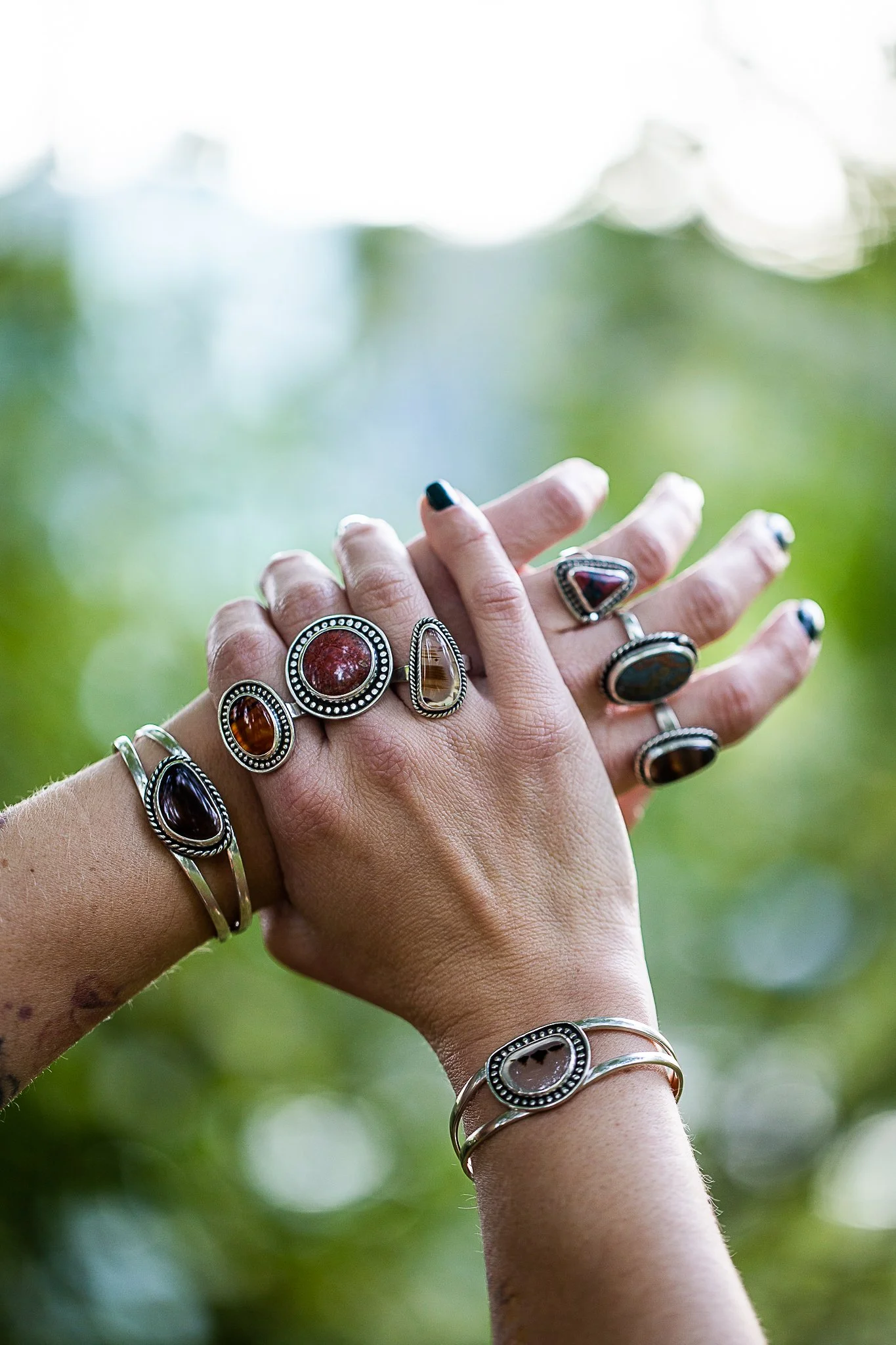 Two hands with multiple rings featuring large stones, set against a blurred outdoor background.