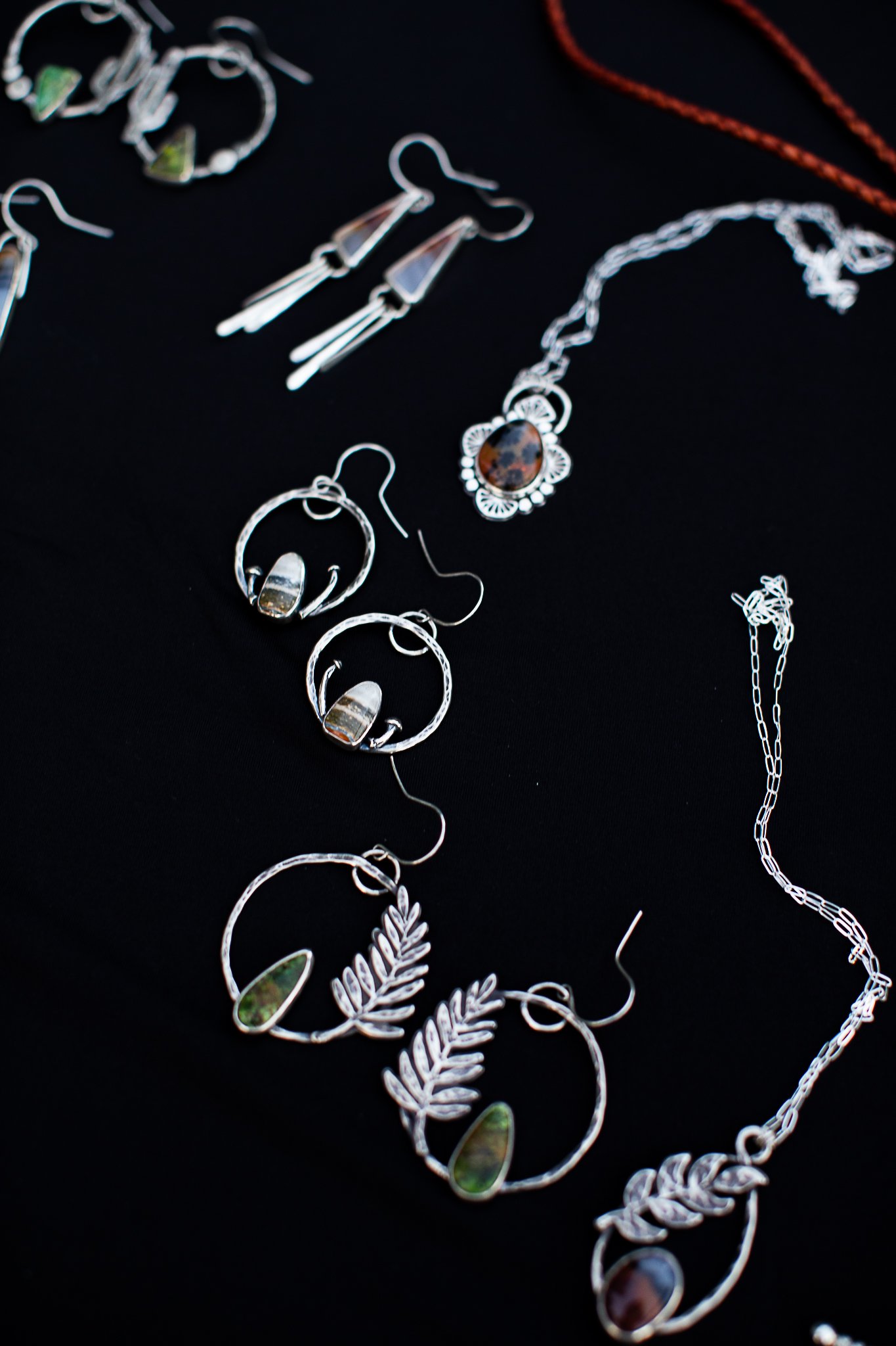 Assorted silver jewelry including earrings, necklaces, and bracelets displayed on a black surface. The jewelry features various stones and decorative elements.