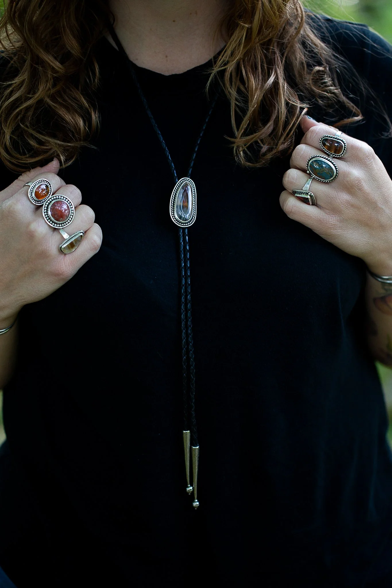 Person wearing multiple rings with gemstone stones and a long necklace with a large pendant, seen from the chest up in a black shirt.