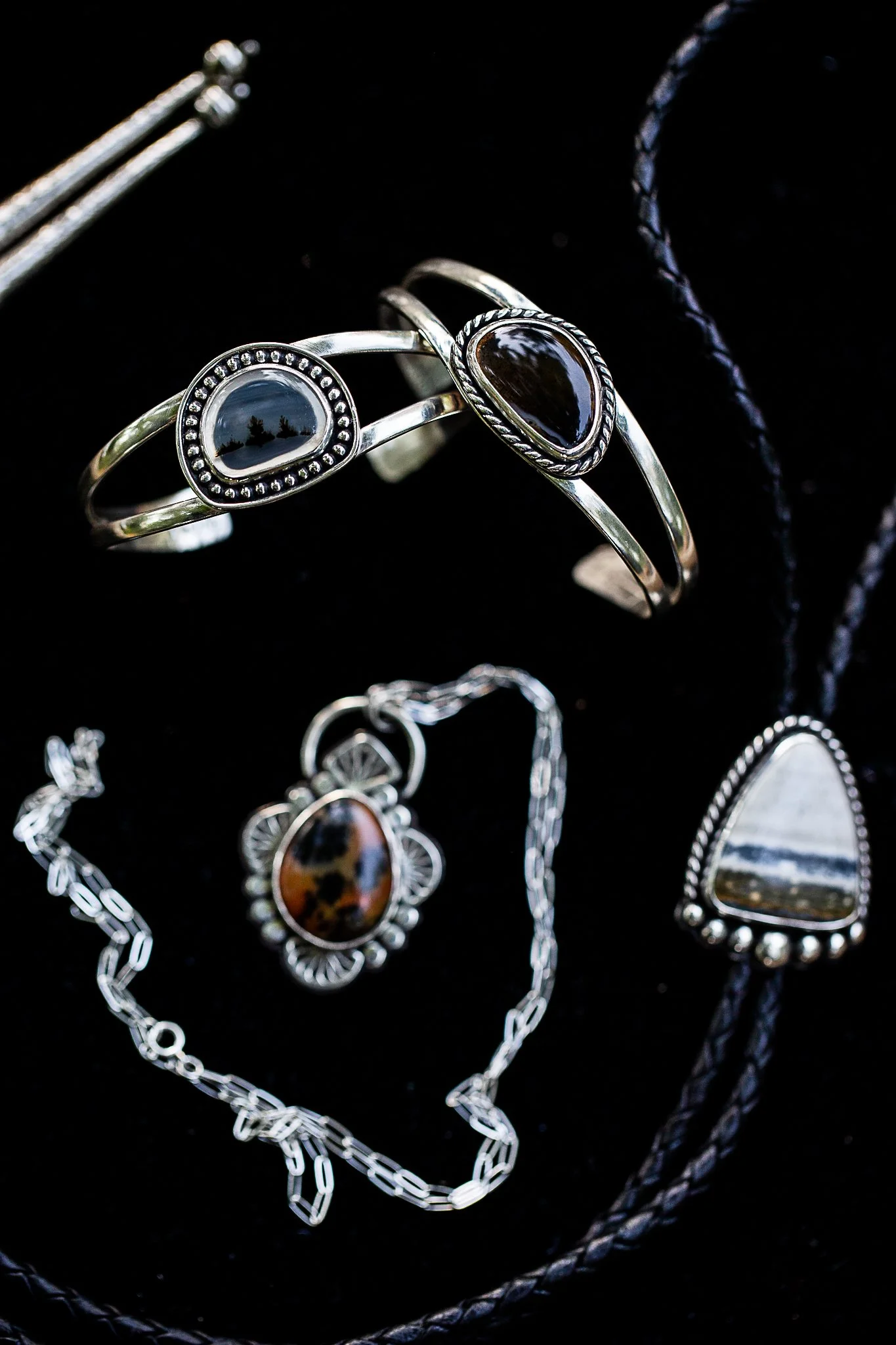 Silver jewelry including rings, a necklace, and a bracelet with black and brown stones displayed on a black surface.