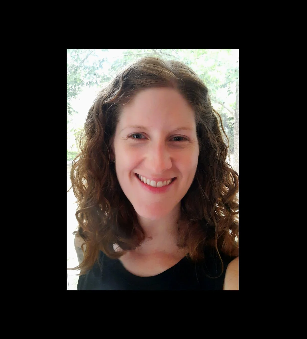 A woman with curly brown hair smiling at the camera, wearing a black top, with a background of greenery visible through a window.