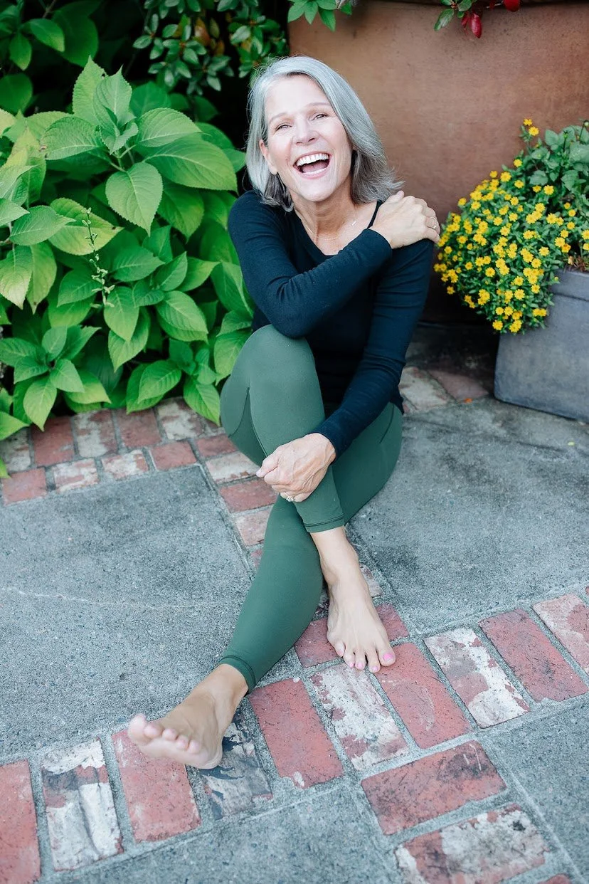 A smiling older woman with gray hair, wearing a black long-sleeve top and green leggings, sitting barefoot on a brick and concrete patio among green plants and yellow flowers.