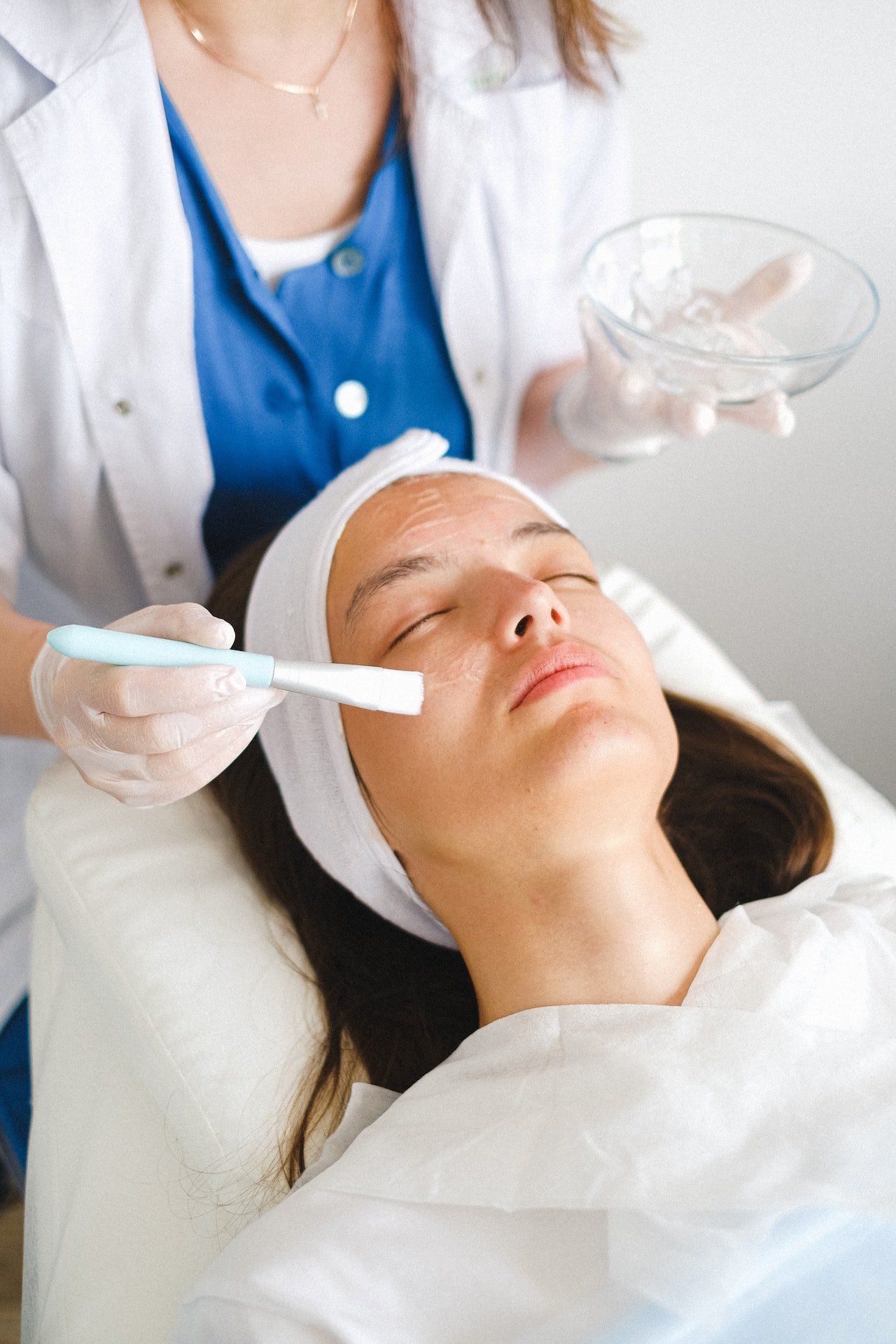 A woman is lying down with her eyes closed while a skincare professional applies a facial treatment using a brush. The woman is wearing a headband and a white gown, and the professional is wearing gloves and a white coat.