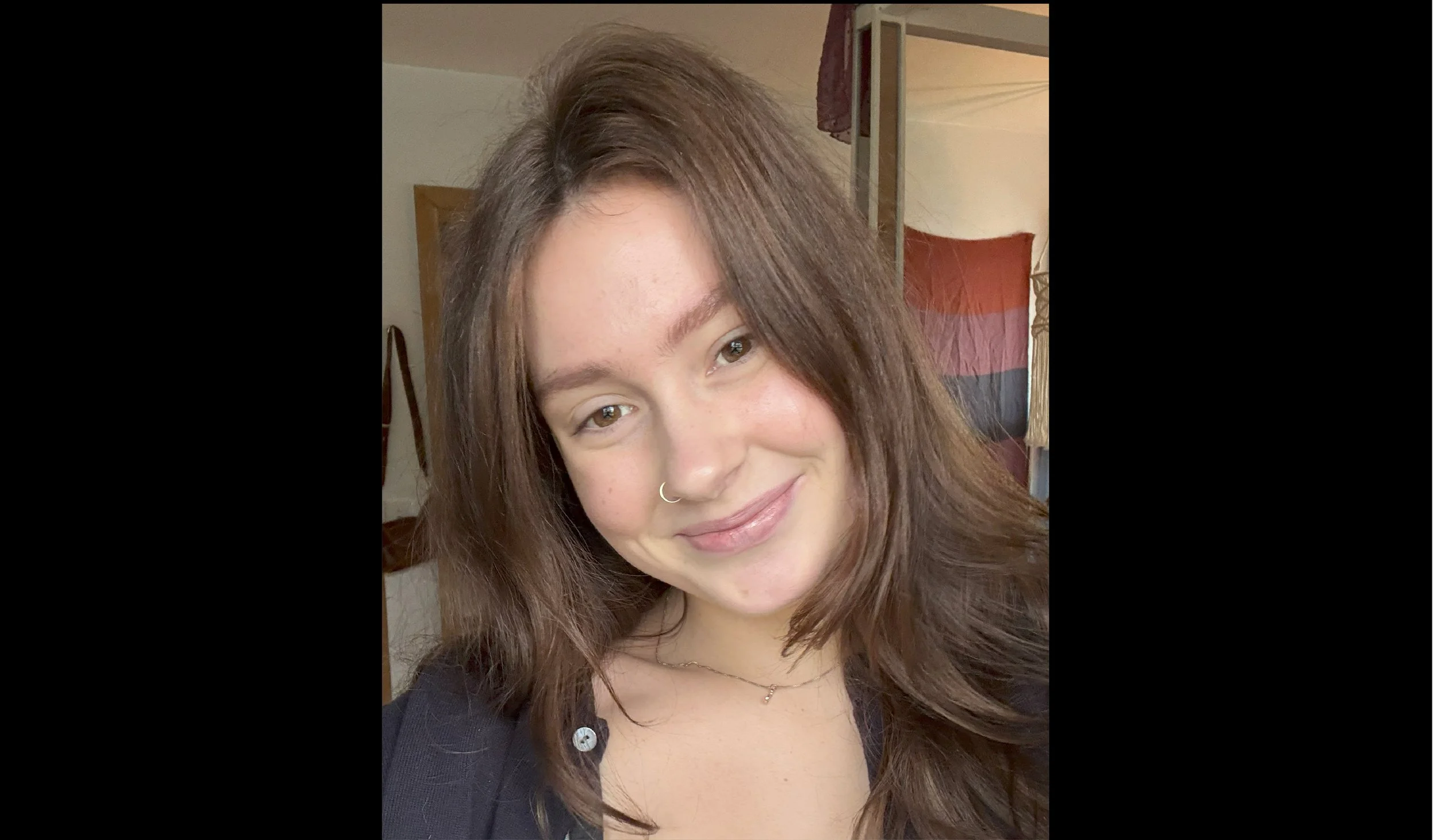 A young woman with light brown, curly hair and a nose ring is smiling at the camera. She is wearing a black top and is indoors, with a background that includes a partially open door and some decorative items.
