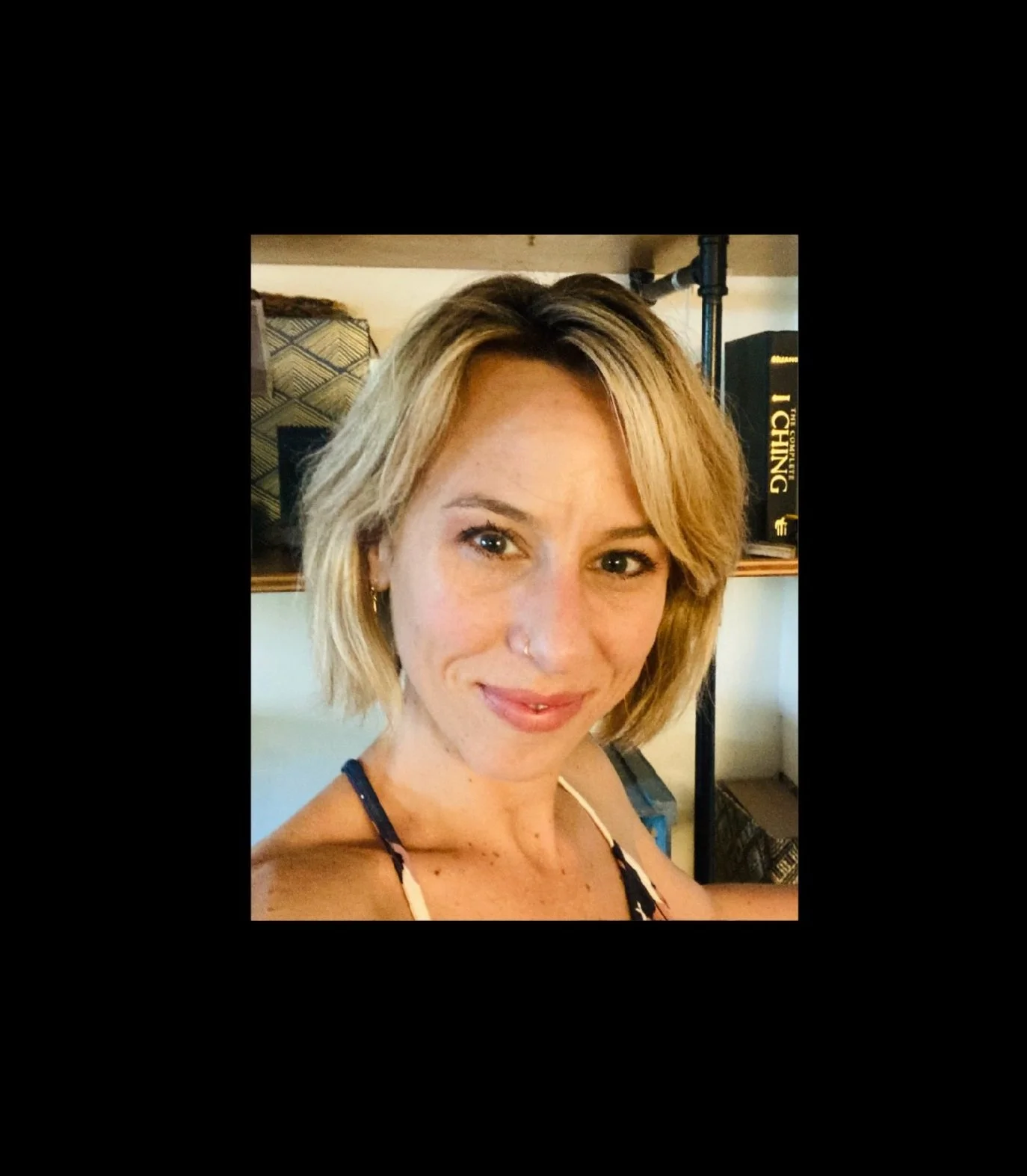 Close-up of a smiling woman with blonde hair, wearing a tank top with thin straps, in a room with books and a black pole in the background.