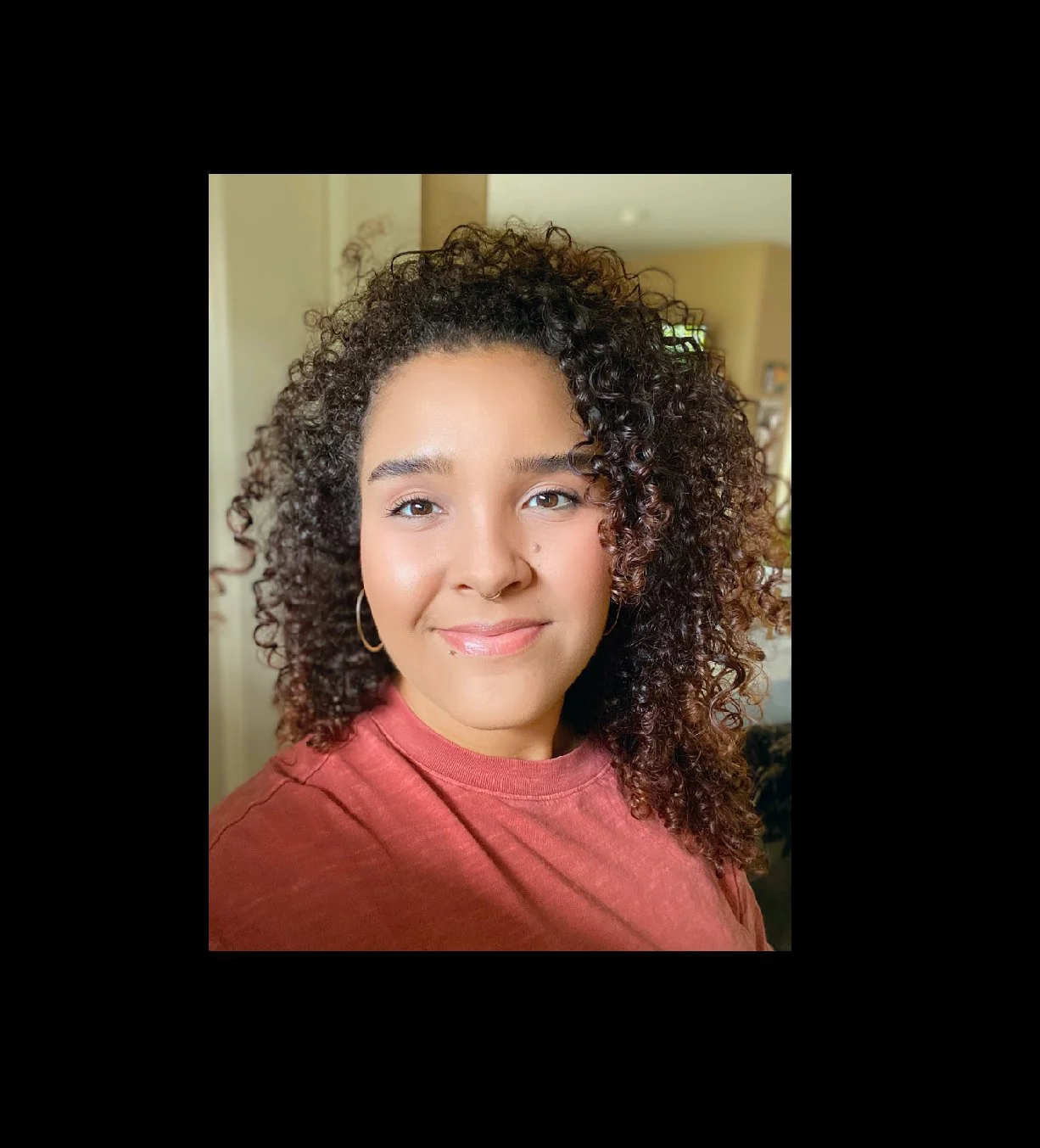 A young woman with curly, dark brown hair and hoop earrings, smiling in a close-up portrait.