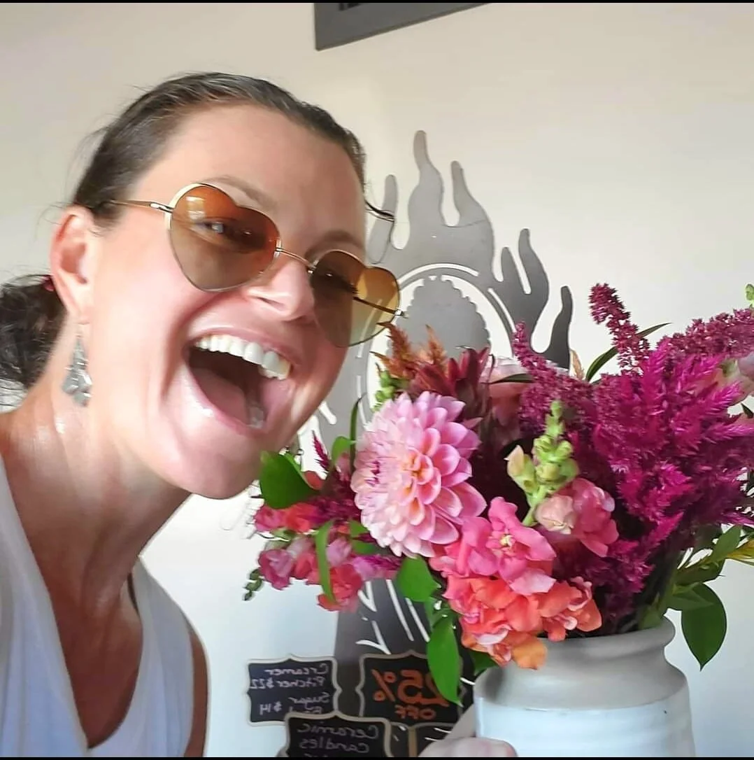 A woman wearing sunglasses and earrings is smiling and holding a white vase with pink and purple flowers.