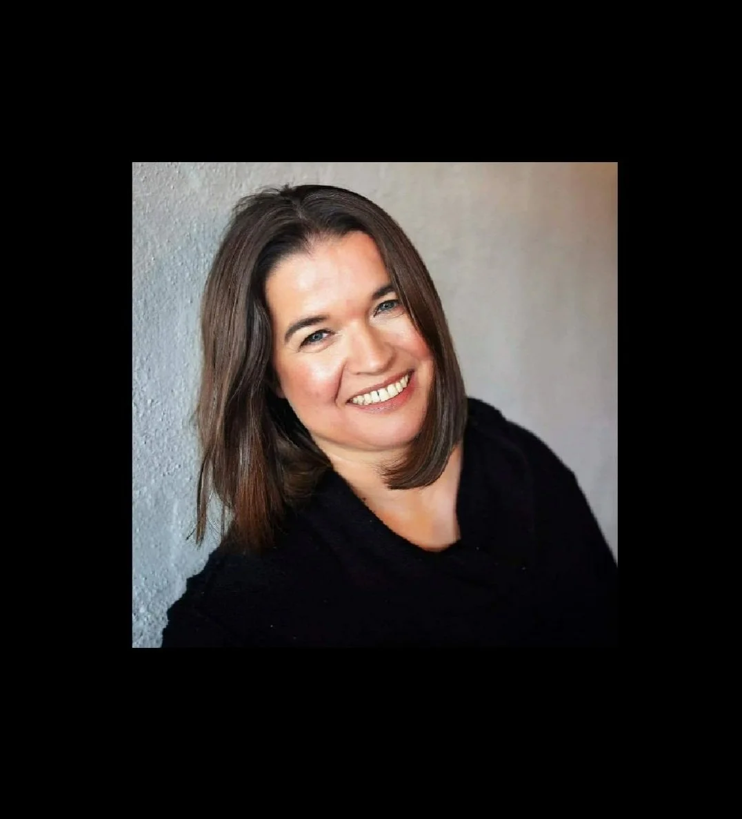 Smiling woman with shoulder-length brown hair, wearing a black top, against a textured light gray wall.