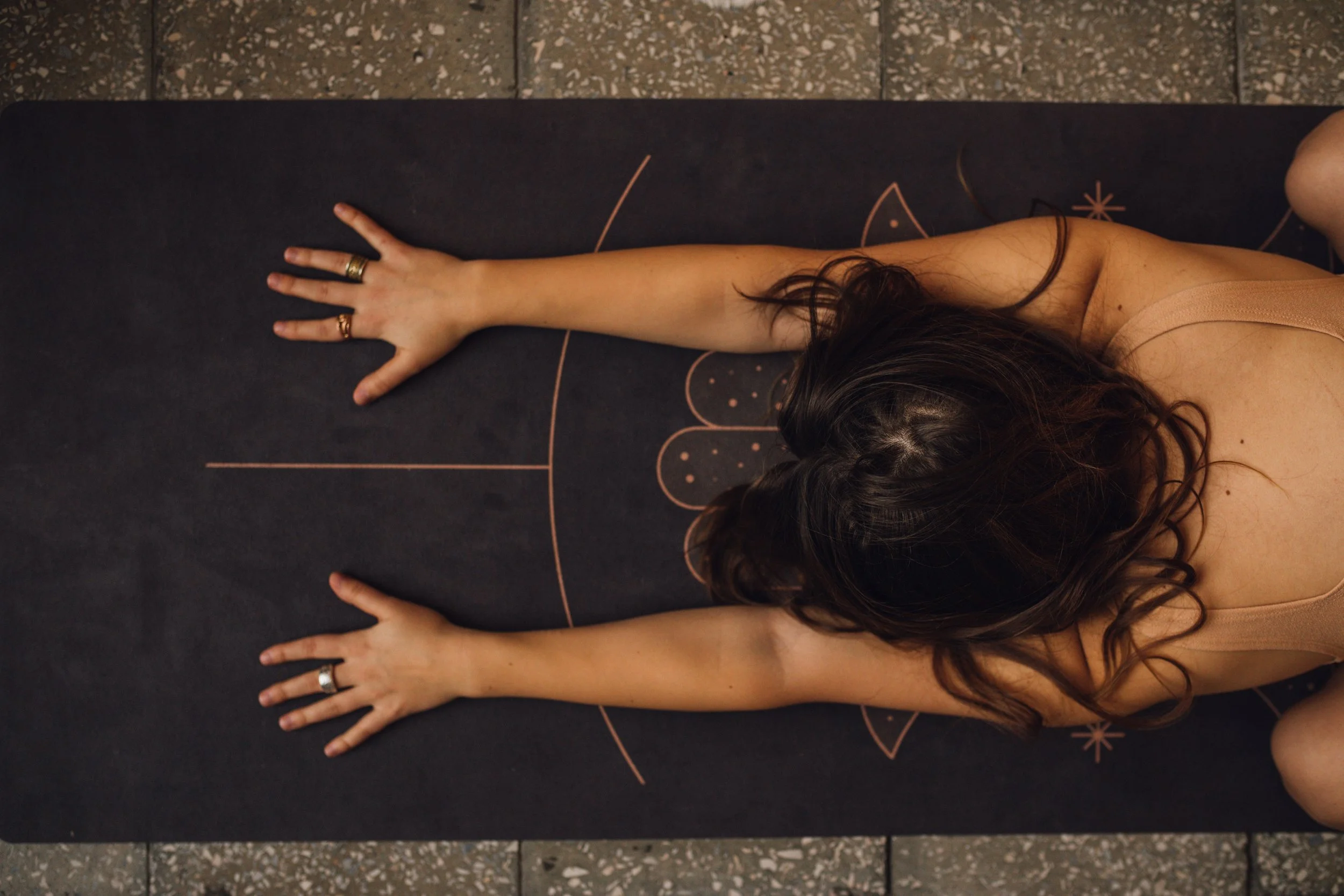 A woman is performing a yoga pose on a black mat with pink and gold designs. She is lying face down, extending her right arm forward with her hand flat on the mat, wearing rings and a beige sleeveless top with her long dark hair flowing.