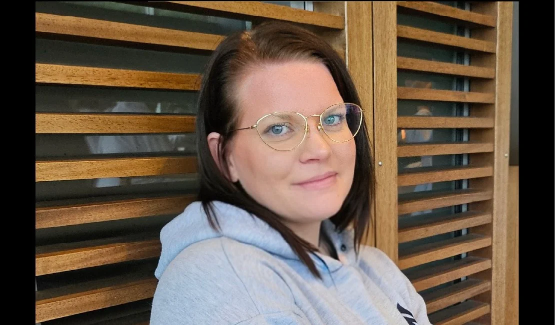 Young woman with glasses and dark brown hair, wearing a light gray hoodie, sitting in front of wooden slats.