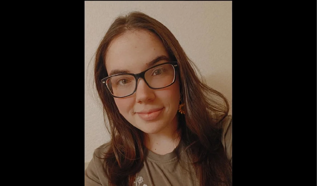 A young woman with long brown hair, glasses, and a septum piercing smiling at the camera, wearing a beige shirt and gold earrings, with a plain light-colored wall background.