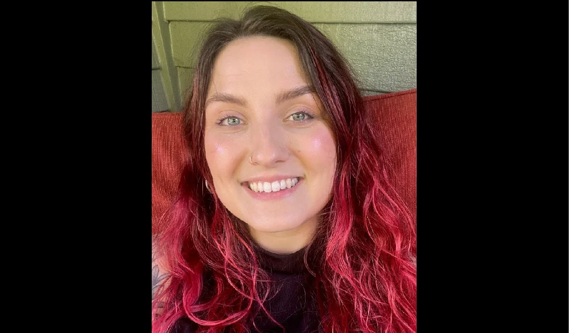 Close-up portrait of a young woman with pink and red wavy hair, smiling, wearing a black top, sitting against a green wall and red cushion.