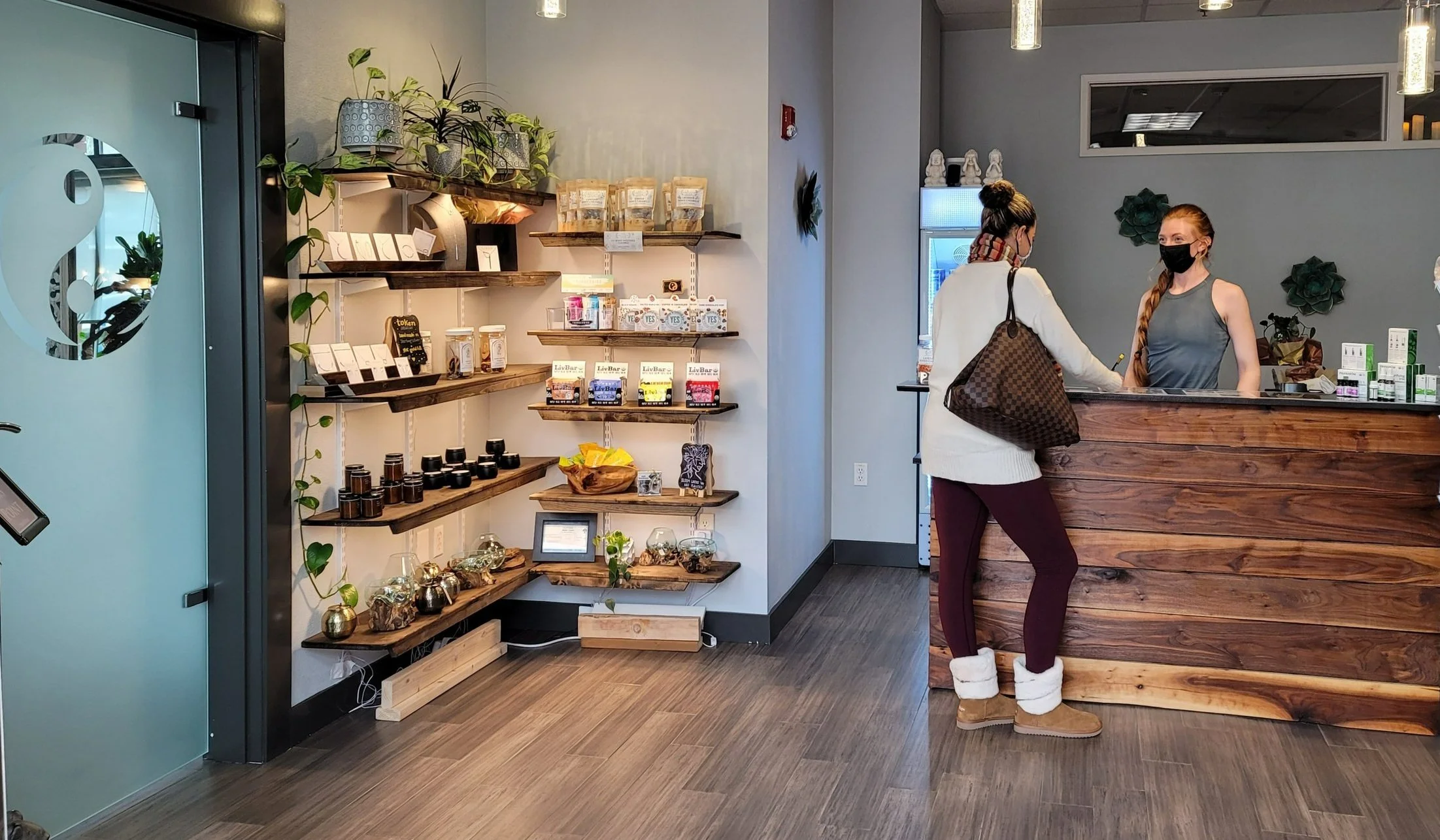 Interior of a wellness or health shop, with a counter on the right where a woman wearing a mask interacts with a staff member behind the counter. Shelves on the left display various health products, potted plants, and decorative items. The setting has a modern, cozy design with wooden accents and subdued lighting.