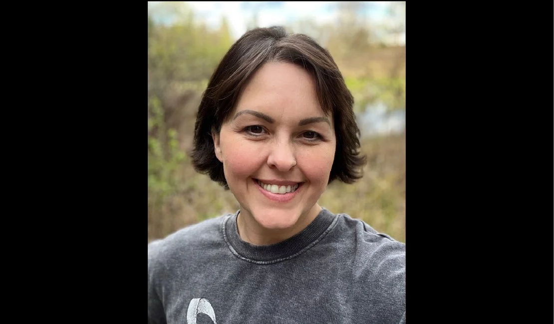 A woman with short dark hair smiling outdoors on a cloudy day, wearing a dark gray T-shirt.