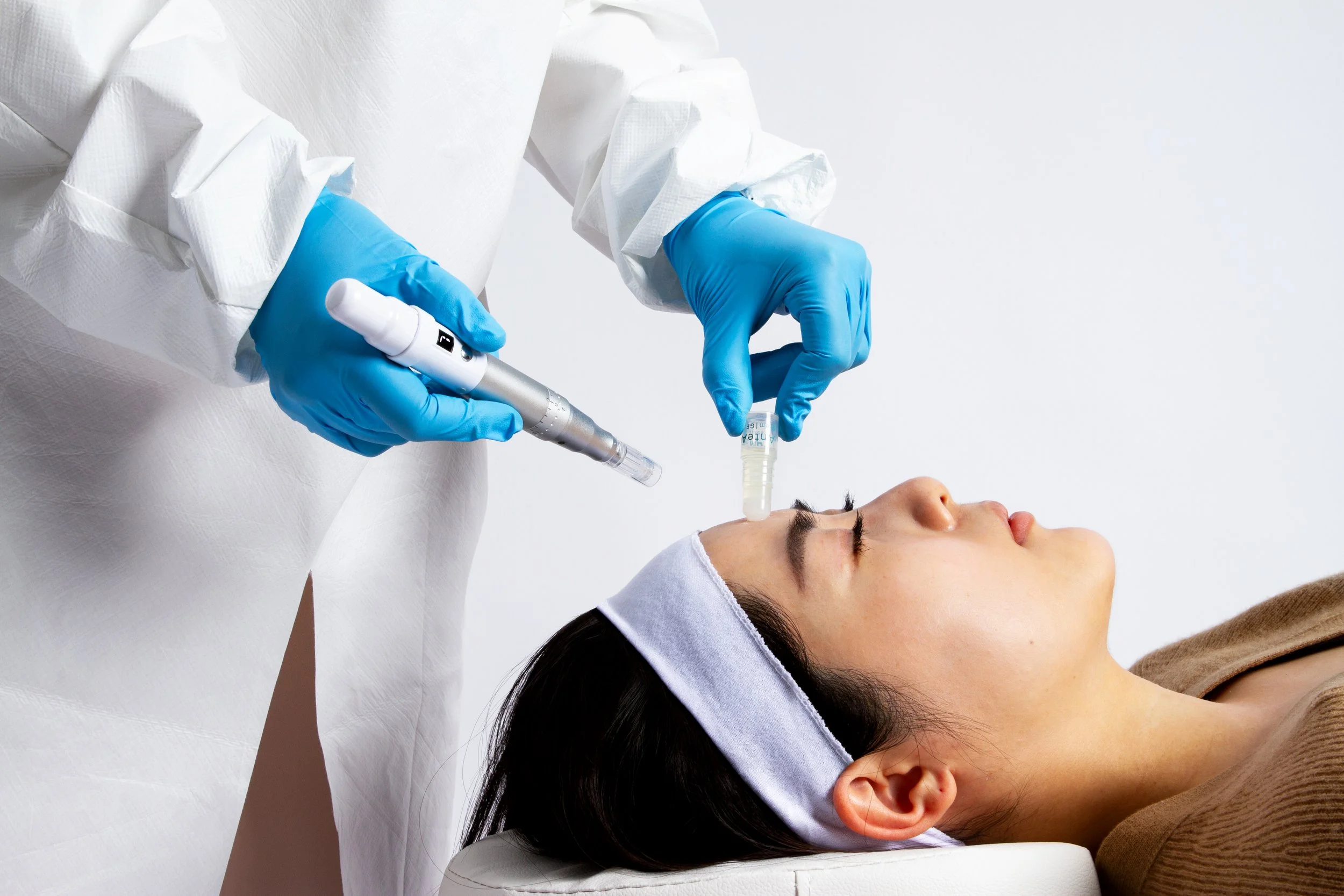 A woman lying down with her eyes closed while a healthcare professional in protective gear performs a procedure with a medical device on her face.