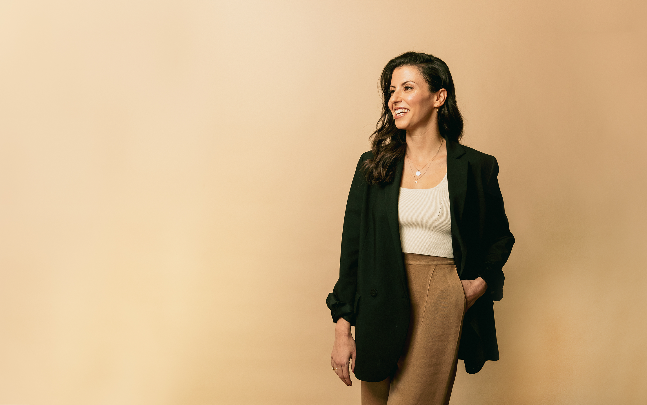A woman with curly brown hair wearing a black blazer, cream top, and brown pants standing against a beige background, smiling and looking to her left.
