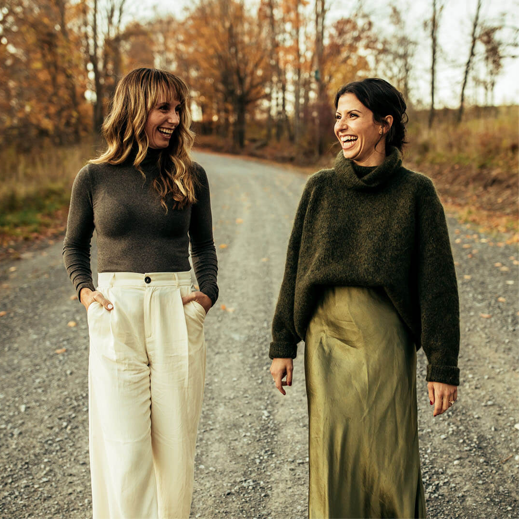 Two women are walking on a gravel path through a forest during fall, smiling and laughing at each other. The woman on the left has wavy blonde hair and is wearing a black turtleneck sweater and high-waisted cream-colored pants. The woman on the right has short dark hair and is wearing a dark green sweater and olive-green pants. The trees in the background are autumn-colored with orange, yellow, and brown leaves.