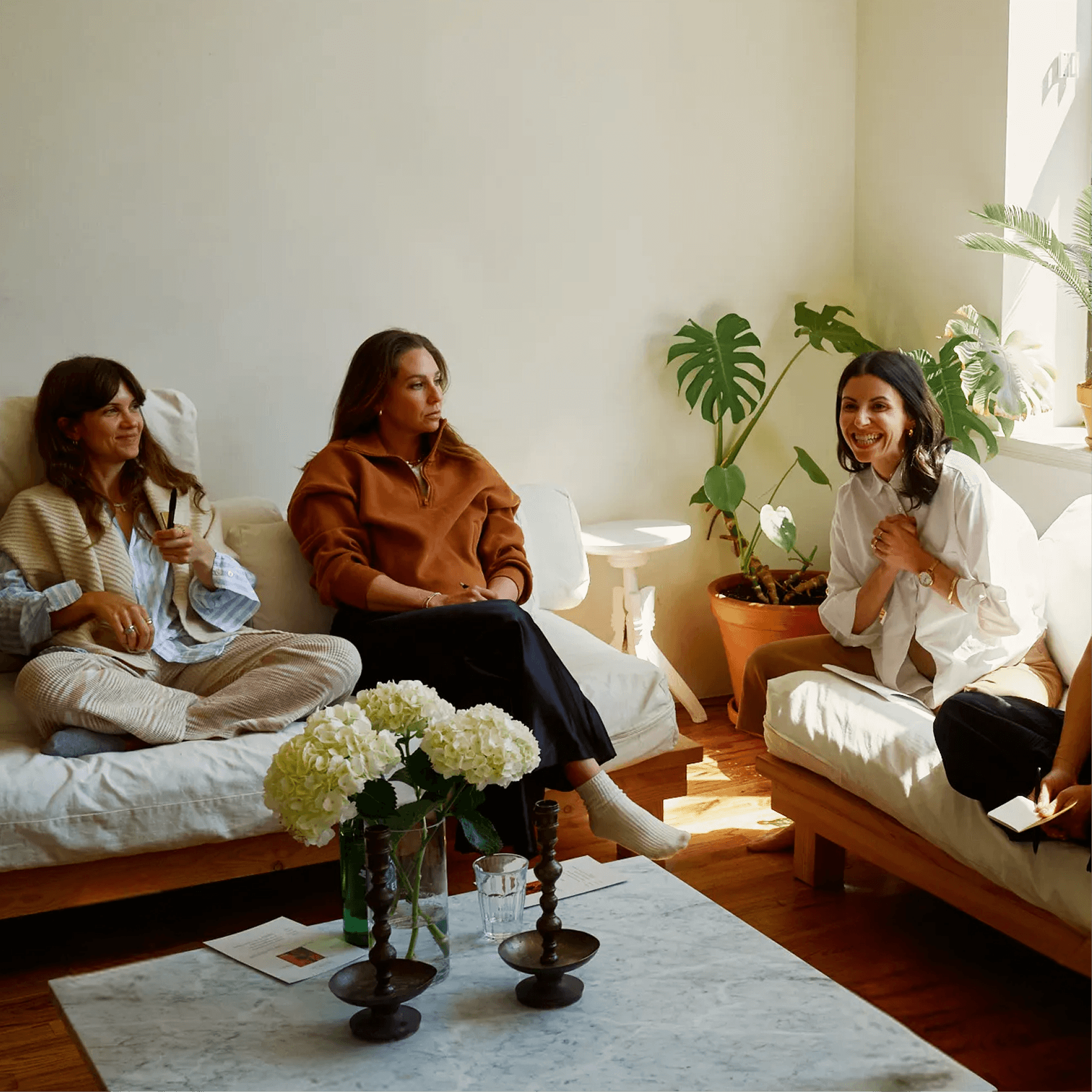 Three women sitting on a sofa and couch in a cozy, sunlit living room with plants, flowers, and reading materials on a coffee table, engaged in conversation.