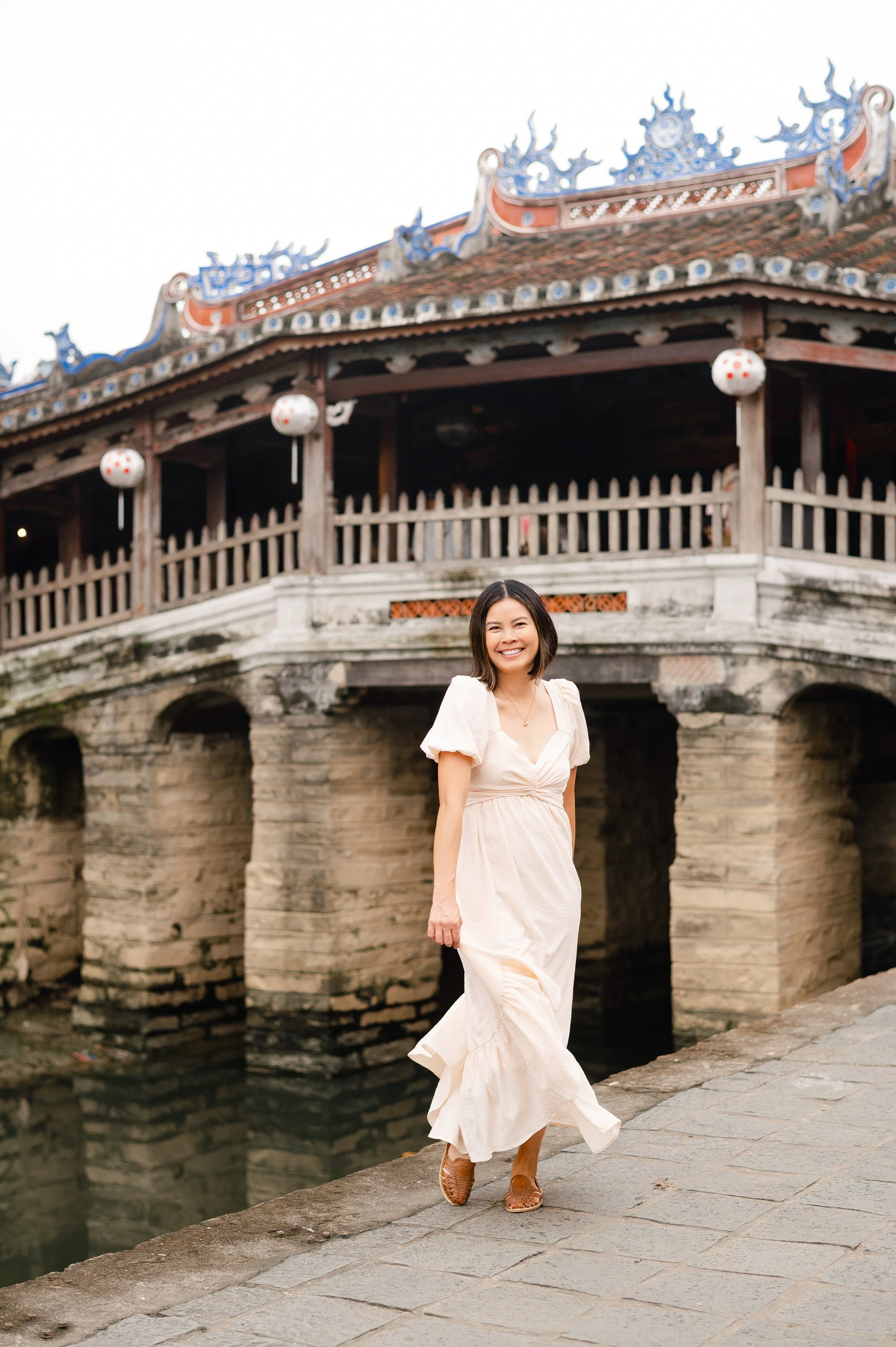 A smiling Carlsbad woman in a white dress walking along a stone pathway near a wooden Asian-style building with a tiled roof and hanging lanterns.