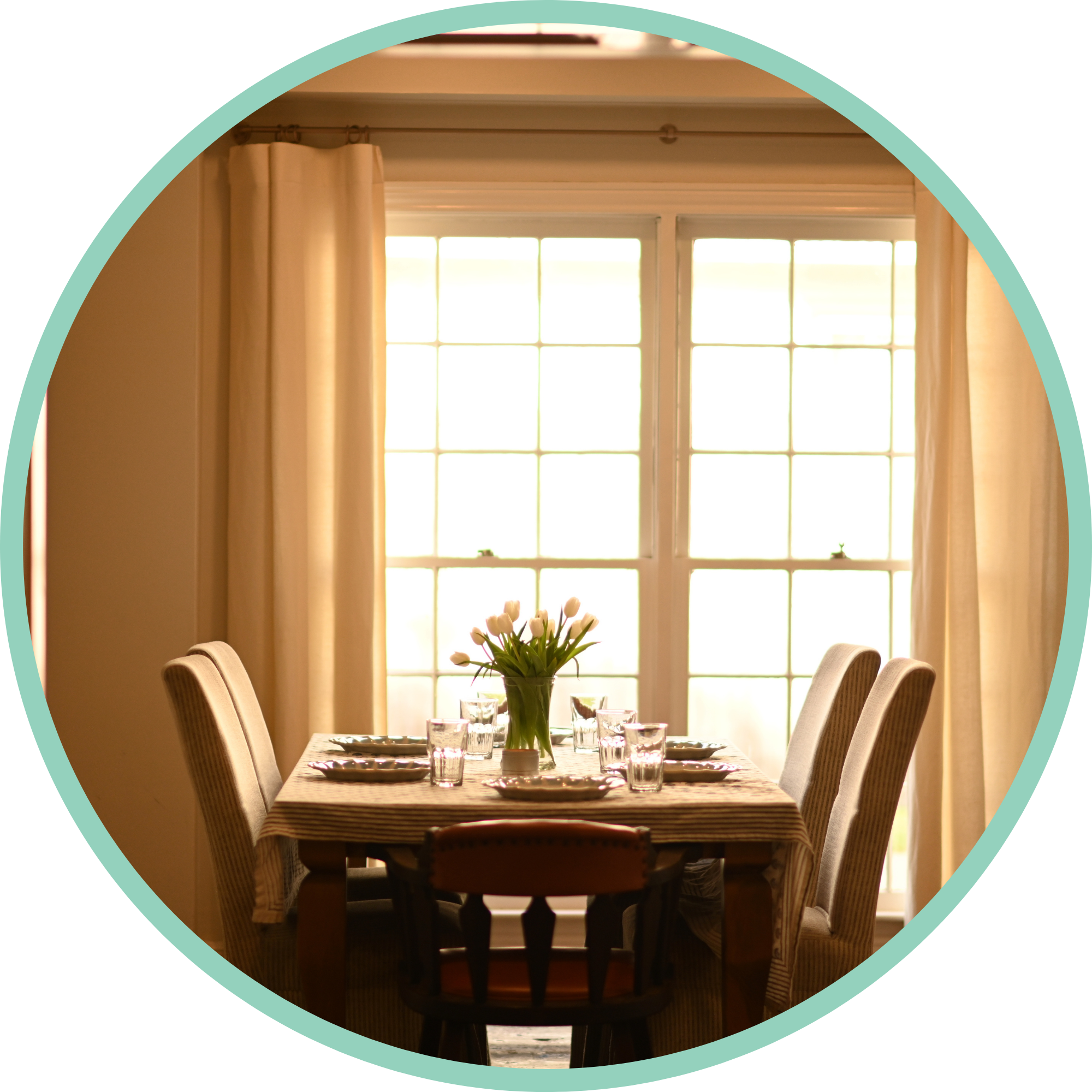 Sunlit dining room with a wooden table set with plates, glasses, and a central vase with white tulips. Beige upholstered chairs surround the table. Large windows with beige curtains let in natural light.