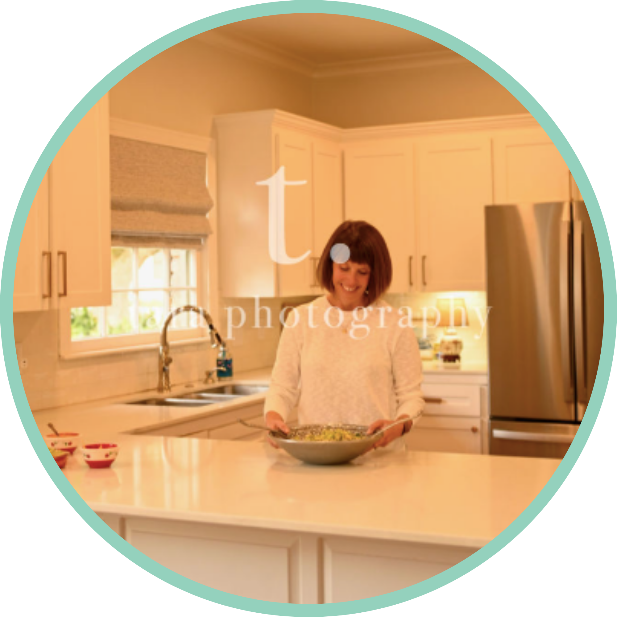 Katie Tasman. A woman with short brown hair smiling in a kitchen. She is holding a baking dish in front of her. The kitchen has white cabinets, a window with a beige shade, and a sink with a faucet. There are small bowls on the counter to her left.