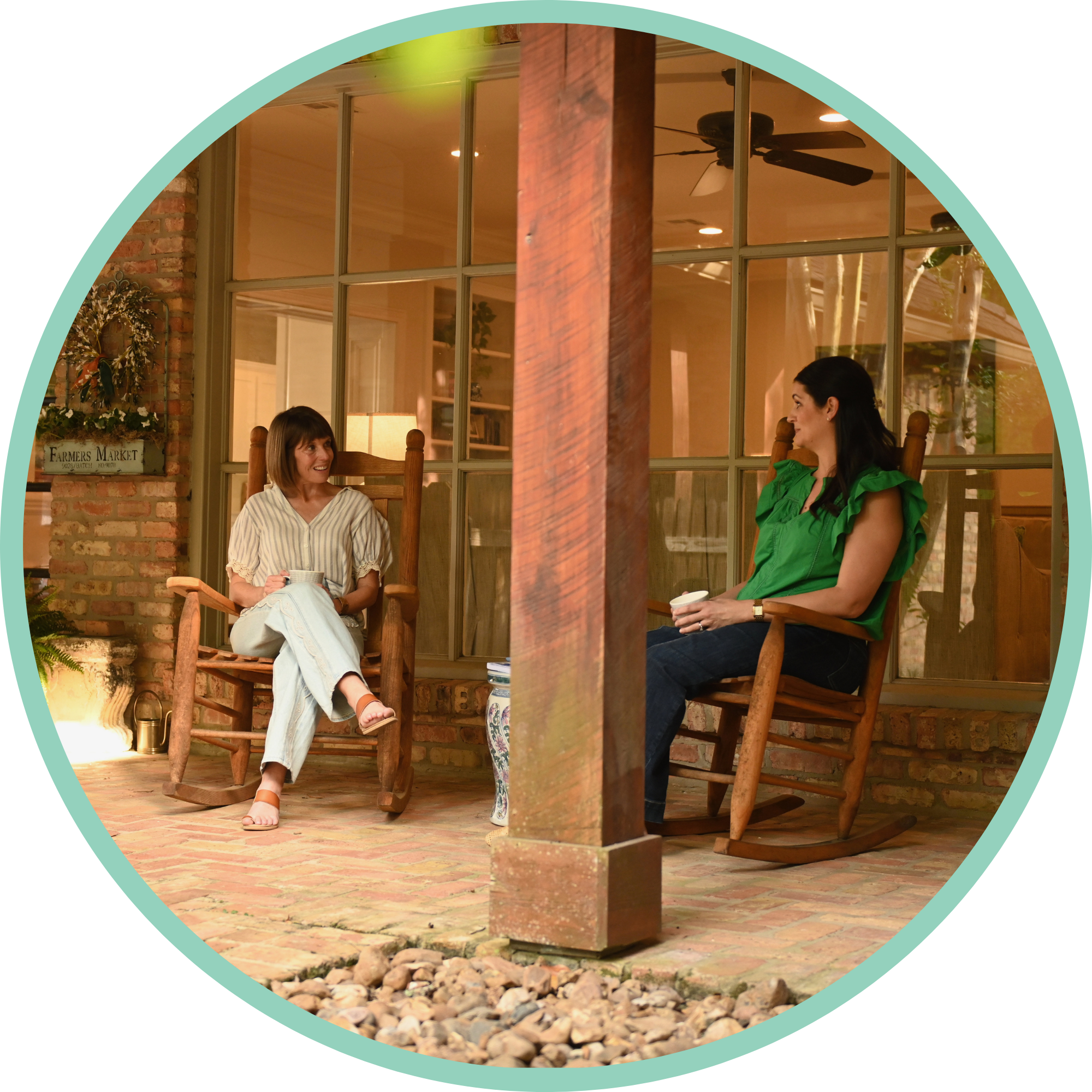 One-on-One Coaching. Two women sitting and chatting on a porch or patio, with a brick wall and large window in the background. One woman is wearing a striped shirt and light jeans, while the other is dressed in a green top, holding a coffee cup.