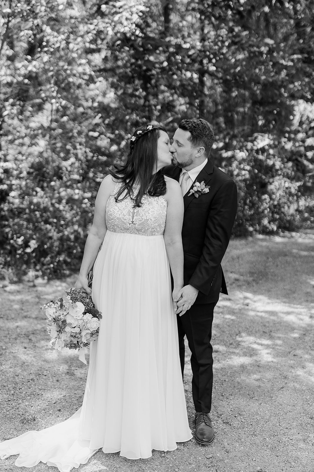 A black and white photograph of a newlywed couple kissing outdoors. The bride is wearing a long, sleeveless wedding dress with lace details and holding a bouquet of flowers. The groom is dressed in a dark suit with a boutonniere. They are holding han