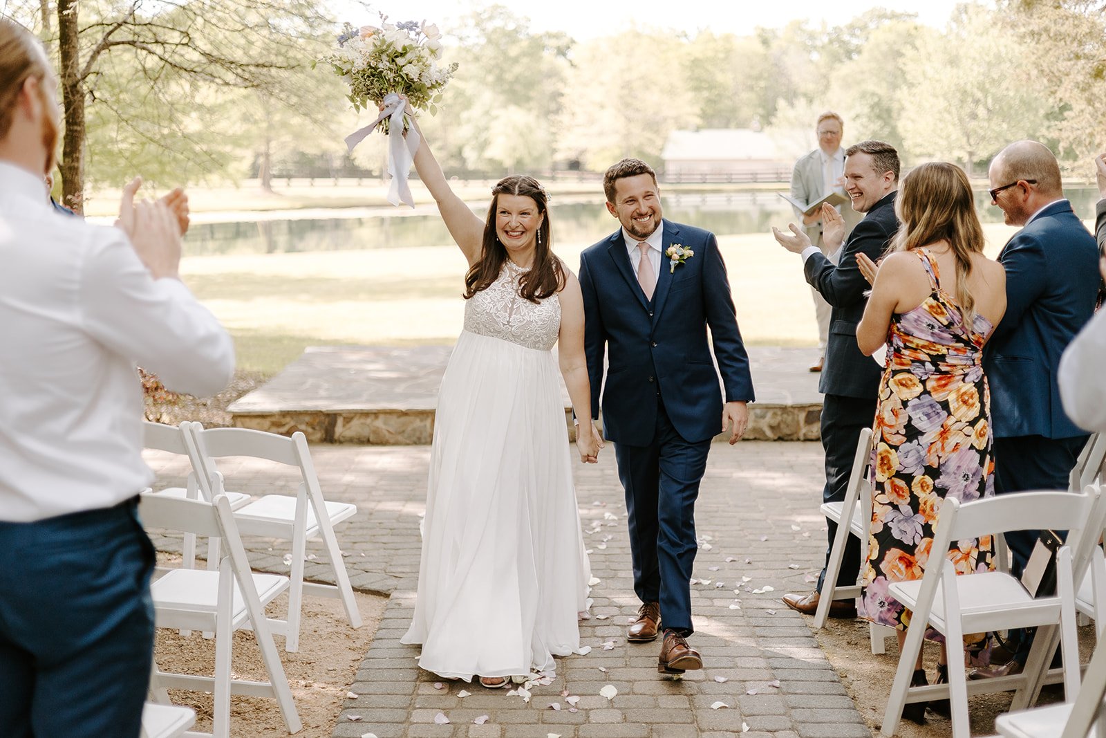 A bride and groom holding hands and walking down the aisle at an outdoor wedding, with guests clapping and smiling.